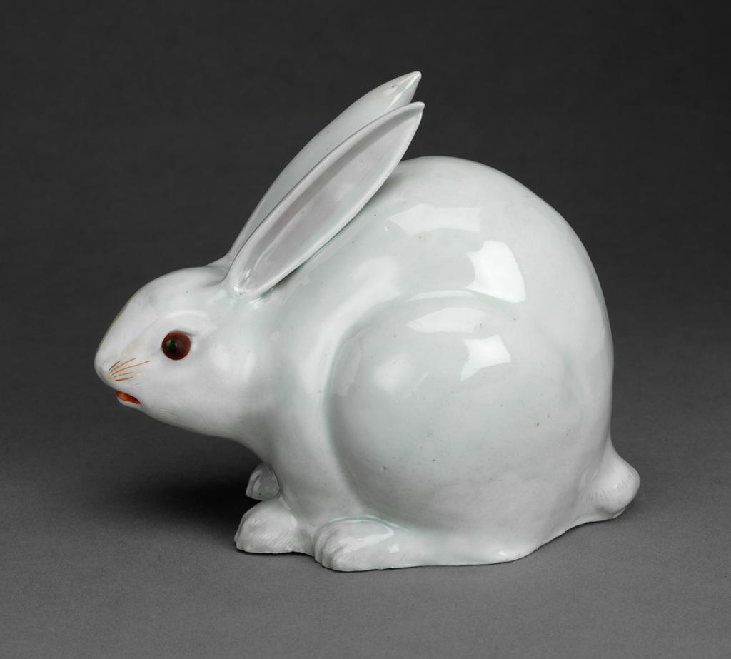 An image of Hare. Hard paste porcelain with greyish glaze and pinkish gold-lustre wash details picked out in red and dark brown and black enamels.  Model of a crouching hare, moulded with hollow interior, the pointed ears, painted with pink-gold lustre on the inside, resting on its arched back, the mouth pierced at either corner and picked out in red enamel; red whiskers; red eyes with dark green pupils; paws and tail with incised details and pinky wash.