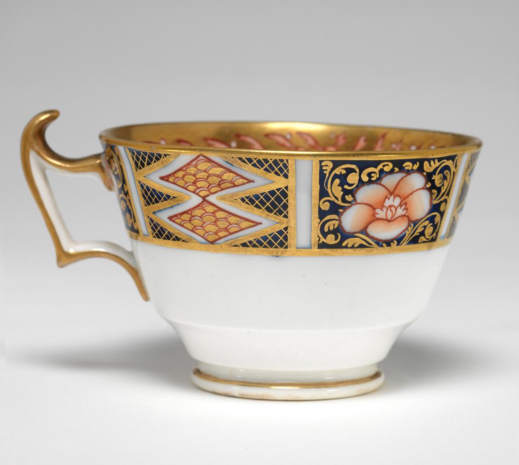 An image of Cup and Saucer/Teacup. New Hall Porcelain Factory, Staffordshire. The cup is of London shape. The circular saucer has deep sloping sides, and stands on a footring. The inside of the cup has a red and black bird on a white area. The rest of the surface is covered with two underglaze blue panels decorated with red and gold flowers, and two white panels decorated with a spray of underglaze blue and green leaves and red flowers, with seven red squares below. Above the panels there are red stylized plants reserved in a gold gound. The exterior is decorated with a border comprising alternating rectangles containing two scale-patterned diamonds reserved in a blue ground, and a red flower reserved in a blue ground. The back of the handle is gilded. The saucer is decorated with the same design as the interior of the cup, with the interior border round the outer edge. Pattern 1368. bone china, painted underglaze in cobalt blue, overglaze in pale green, iron-red, a little black, and gold. Height, cup, 6 cm, width, cup, 10.6 cm, diameter, cup, 8.7 cm, height, saucer, 2.8 cm, diameter, saucer, 14 cm, circa 1813-1818. Imari Style.