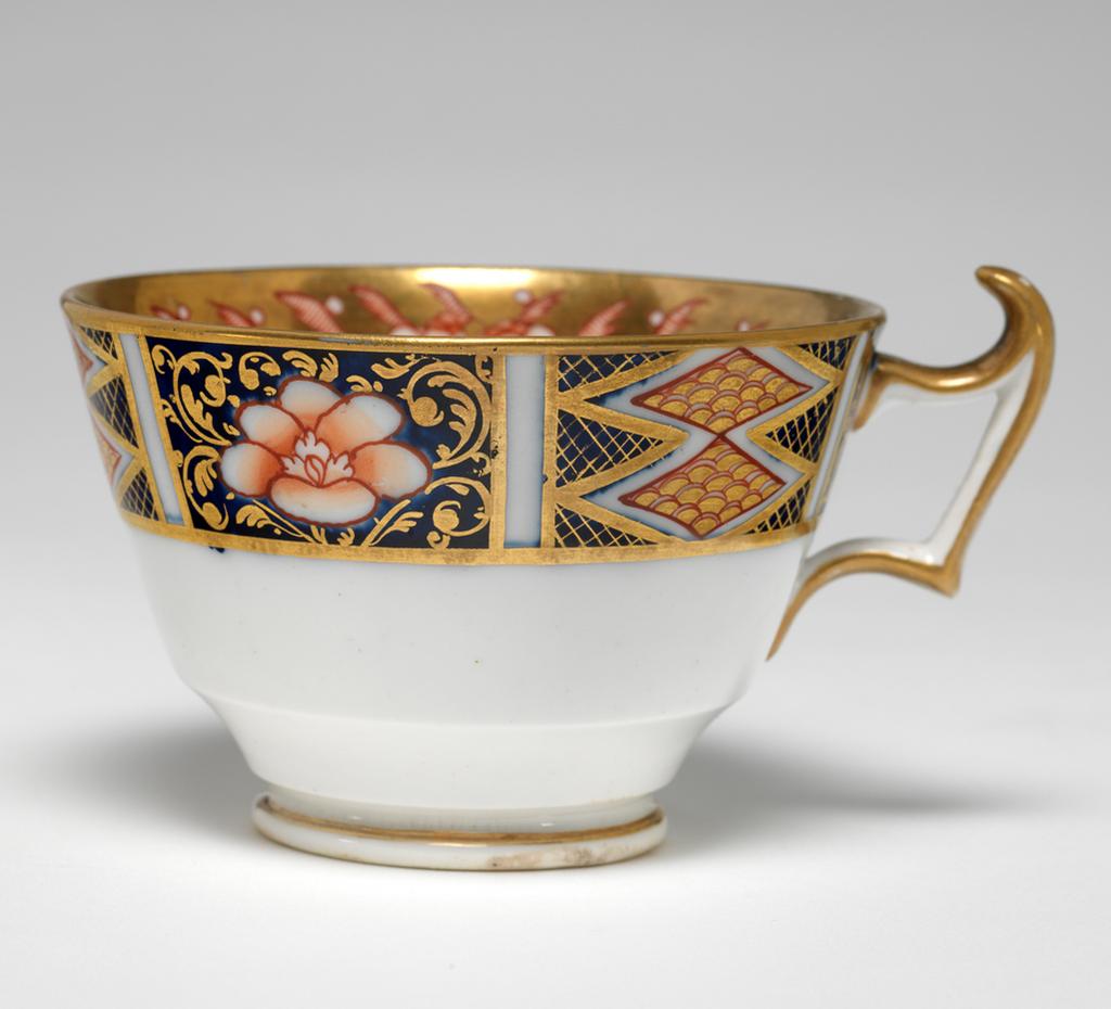 An image of Cup and Saucer/Teacup. New Hall Porcelain Factory, Staffordshire. The cup is of London shape. The circular saucer has deep sloping sides, and stands on a footring. The inside of the cup has a red and black bird on a white area. The rest of the surface is covered with two underglaze blue panels decorated with red and gold flowers, and two white panels decorated with a spray of underglaze blue and green leaves and red flowers, with seven red squares below. Above the panels there are red stylized plants reserved in a gold gound. The exterior is decorated with a border comprising alternating rectangles containing two scale-patterned diamonds reserved in a blue ground, and a red flower reserved in a blue ground. The back of the handle is gilded. The saucer is decorated with the same design as the interior of the cup, with the interior border round the outer edge. Pattern 1368. bone china, painted underglaze in cobalt blue, overglaze in pale green, iron-red, a little black, and gold. Height, cup, 6 cm, width, cup, 10.6 cm, diameter, cup, 8.7 cm, height, saucer, 2.8 cm, diameter, saucer, 14 cm, circa 1813-1818. Imari Style.