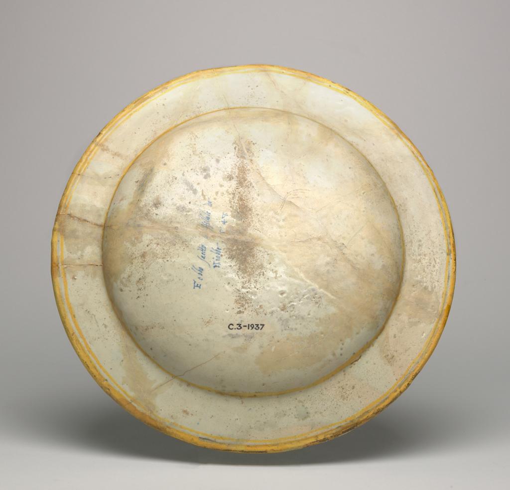 An image of Maiolica dish. Apollo slaying the Sons of Niobe. Guido di Merlino, workshop, possibly (Italy, The Marches, Urbino). Polidoro da Caravaggio (Polidoro Caldara), painter, after (Italian, 1490/1500-1543). Shape approximately 64 with plain rim. Circular with almost flat rim, and wide shallow well; warped in firing. Apollo stands on the left, firing his bow towards the stampeding horses of the sons of Niobe, five of whom are wholly or partly visible. Behind Apollo is the head of an old man. Further back to left there is a group of trees on either side of a rock, and to right, the citadel of Cadmus and two trees. In the distance there is a landscape with a mountain. A black band and a wider yellow band encircle the outer edge. The back is inscribed in the middle in blue: `Febbo saetto li ffillole de/Niobbe .548' (Phoebus shoots the sons of/Niobe). Earthenware, tin-glazed overall; the reverse pale beige. Painted in blue, green, yellow, orange, stone, brown, manganese-purple, black, and white, height 4 cm, diameter, rim, 42.8 cm, dated 1548. Renaissance. Production Note: The design was derived from the facade of the Palazzo Milesi, via della Maschera d'Oro, Rome, which Polidoro da Caravaggio (1492-1543) completed before the Sack of Rome in 1527. It was the most celebrated of his facades and was the last of those listed by Vasari. The facade has deteriorated so that only the faintest shadows of its former splendour remain. But the whole scheme can be seen in Enrico Maccari's Graffiti e chiaroscuri esistenti nell'esterno delle case di Roma, undated but published in 1876. The frieze illustrating the story of Niobe ran from left to right along the facade above the doorways and below the first-floor windows. No print of this facade is known before 1548 so presumably the painter of the dish had access to drawings made in Rome.