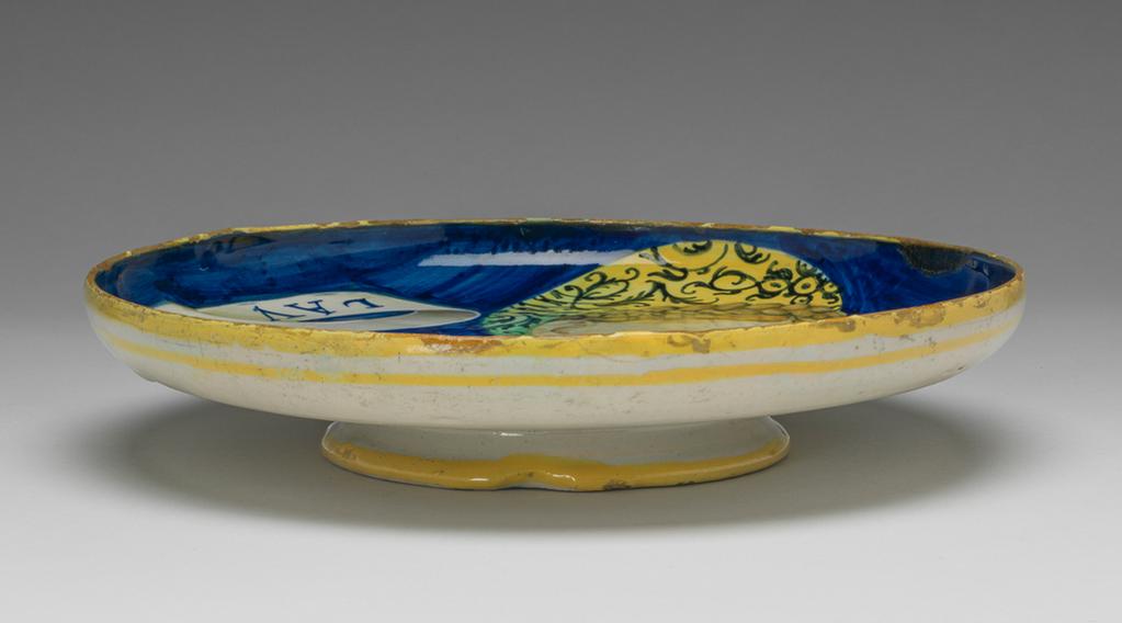 An image of Maiolica. Shallow bowl on low foot. Unidentified potter. This type of dish is traditionally attributed to Castel Durante, but could have been made in Urbino. A bust of a young woman, three-quarters to right, her gaze cast downwards to her right. Her blonde hair is dressed in a plait over the crown of her head. She wears a green and yellow turban embroidered with black scrolling foliage; two gold chains and a rope of beads; and a green dress with a yellow chemise showing on the shoulders. A scroll inscribed `LAVRA/BELLA' in blue projects on either side of her head. The background is blue. On the back are four concentric yellow bands: on the edge of the foot, just beyond the junction of the foot and bowl, and two round the rim. Pale buff earthenware, tin-glazed overall. Painted in blue, green, yellow, orange, cream, and black. Height, whole, 4.7 cm, diameter, whole, 22.7 cm, circa 1525-1535. Renaissance.