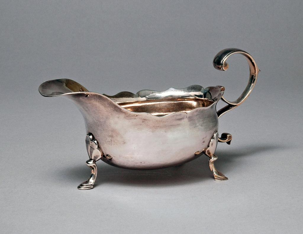 An image of Sauce boat. H?, silversmith, Ireland, Dublin. Oval with wavy edged rim; the sides undecorated; standing on three cast legs with trefoil capitals, knuckles, and hoof feet; with double scroll flying handle, the base scratch engraved with the initials ‘MM’ and ‘5:4½’ for 5 oz 4 ½ dwt. Silver, height, to top of handle, 8.2 cm, width, from lip to handle, 16 cm, weight, overall, 170 g, circa 1760.