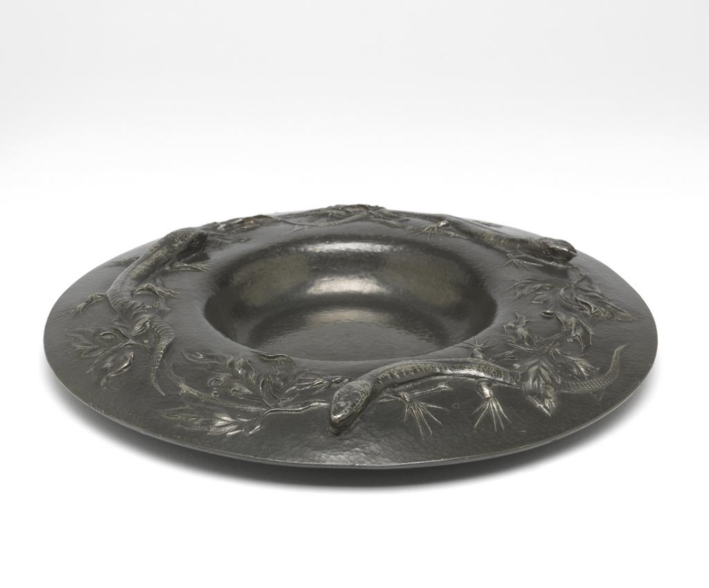 An image of Plate. Marks, Gilbert Leigh (British, 1861-1905). Decorated in relief with three lizards amid foliage. Circular with wide, downward curving rim, and small, deep well. Pewter, height, 2.8, cm, diameter, 23.5, cm, dated 1902. Arts and Crafts. Acquisition Credit: Given by Mrs J. Hull Grundy.