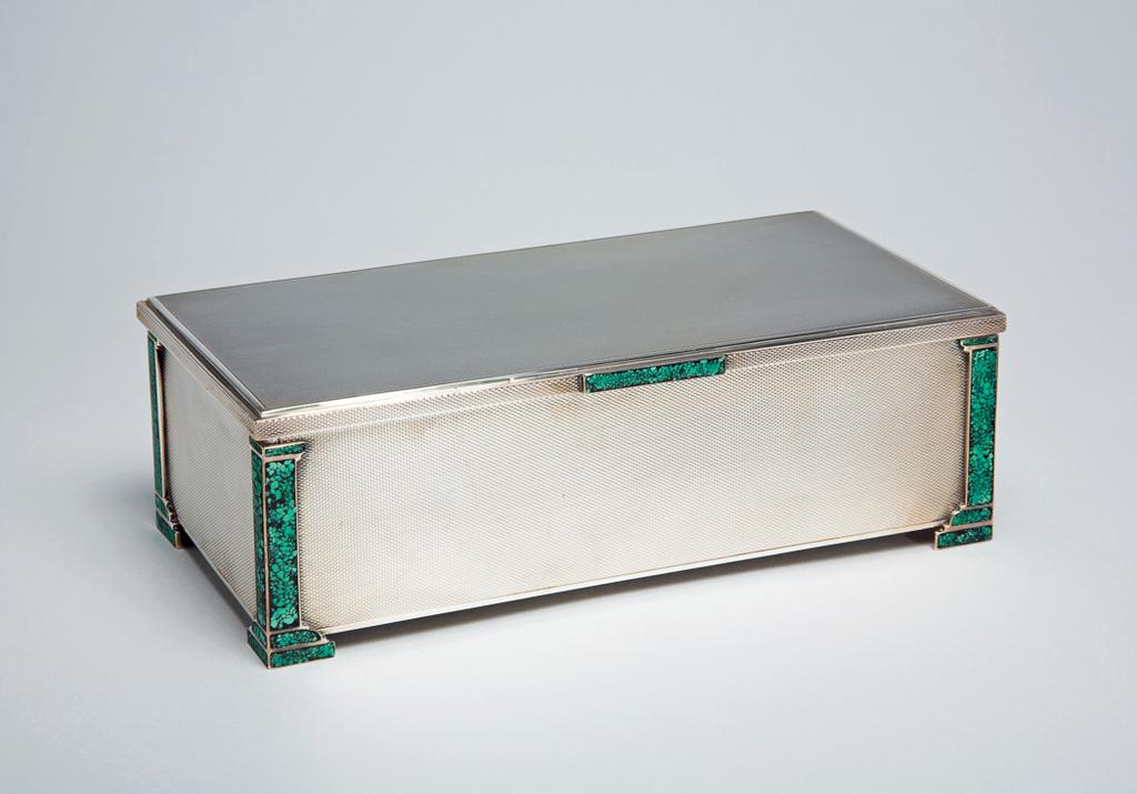 An image of Cigarette box. Mappin & Webb Ltd. Rectangular. The sides are engine-turned with a fine mesh pattern, and at each corner have a mount in the shape of a 'malachite' enamelled column, the bases of which serve as feet. The lid is decorated with a similar mesh pattern and has a stepped edge and rectangular 'malachite' thumbpiede at the centre front. The underside of the lid is inscribed. The wooden interior is divided into two by a fixed wooden wall and has another which is moveable. The base of the box is covered in black leather. Silver, seamed, engine turned, enamelled and engraved. Height, to top of lid, 6 cm, width, overall, 18.5 cm, depth, overall, 9.6 cm, 1934-1935. London, England. Art Deco.