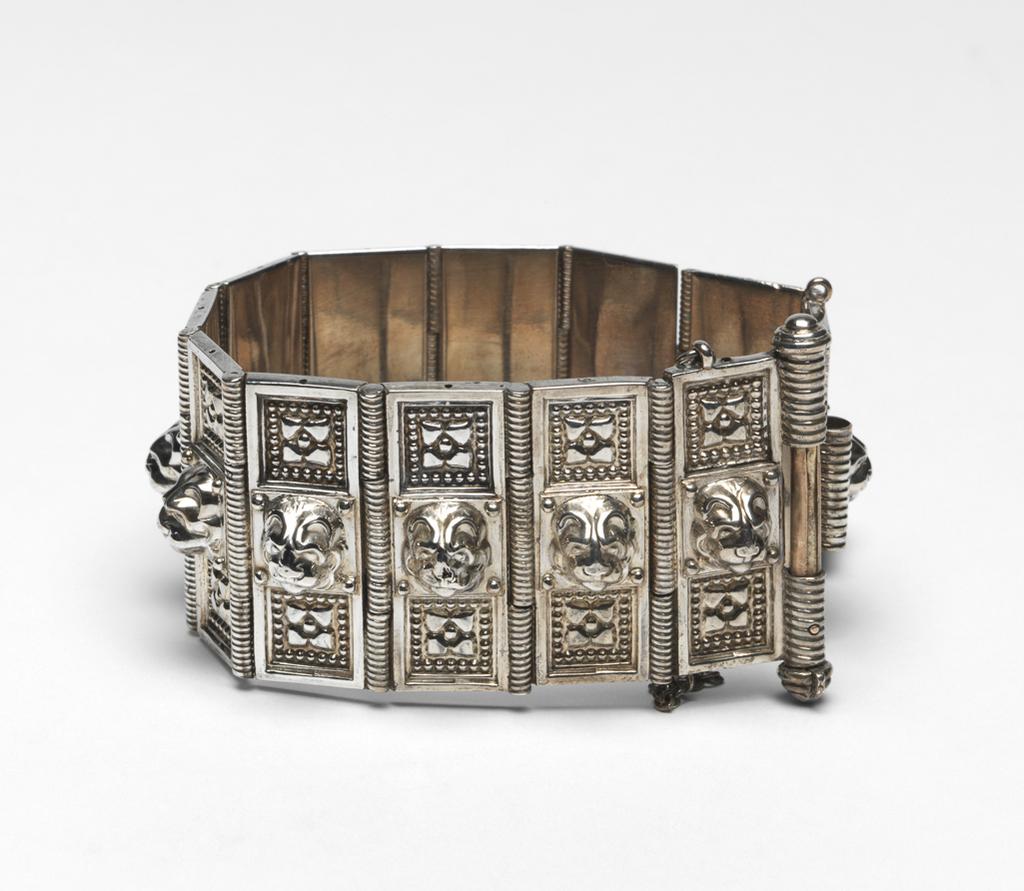 An image of Jewellery. Bracelet. Giuliano, Carlo (Italian, 1831-1895). Composed of twelve hinged panels, each decorated with a lion's head in relief with a four-petalled flower head in a sunk beaded square above and below. The fastening is by means of a bolt passing through three sections of a tube, two on one end of the bracelet and one in the middle of the other. In recent case (A). Rectangular, of dark blue leather with a gilt line round the edge of the lid, the lower part of the interior lined with royal blue velvet, the lid with white satin, printed in gold with the Royal Arms and 'BY APPOINTMENT TO/H M THE QUEEN/JEWELLERS/Wartski/14 GRAFTON ST/LONDON WI. Silver, cast and chased, height, whole, 4.3 cm, length, whole, 18.5 cm, circa 1863-1895. Acquisition Credit: Given by Mrs J. Hull Grundy.