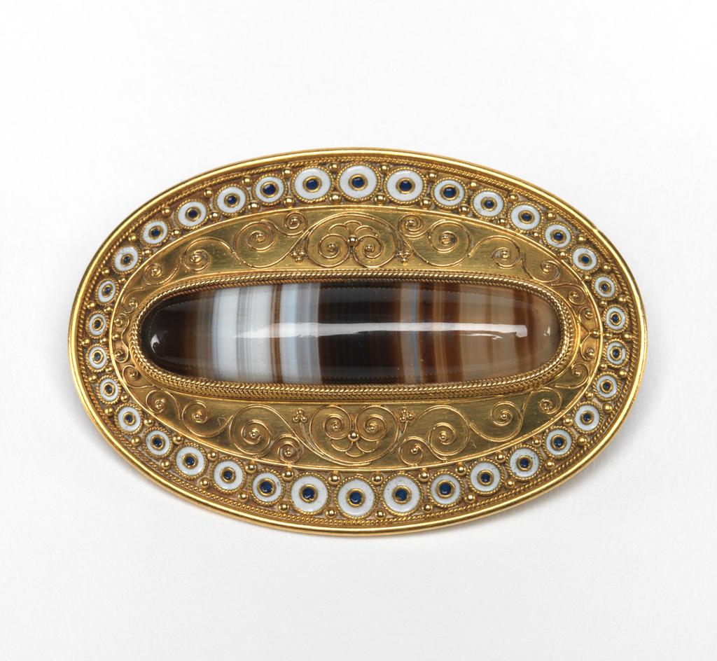 An image of Jewellery. Brooch. Phillips, Robert (British, d.1881). Of slightly convex, oval shield shape with a pin fastening across the back. Inscribed ‘M.P.K/Christmas, 1865’ and 'K.J.K.'. Gold, set with a banded agate and decorated with filigree, granulation, and black and white enamel. Height, whole, 4.8, cm, width, whole, 3.2, cm, dated 1865. Production Note: The brooch may have been a pendant, the present clasp forming the loop at the top. Acquisition Credit: Given by Mrs J. Hull Grundy.