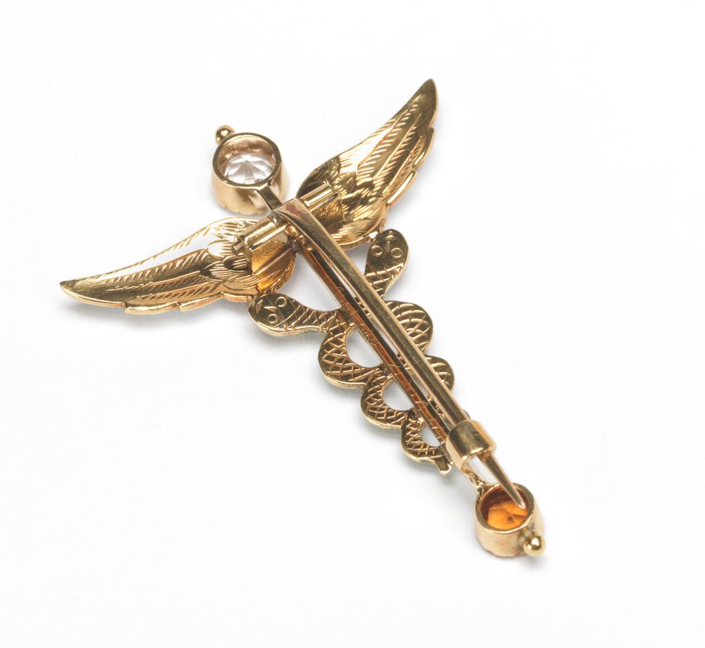 An image of Jewellery. Caduceus Brooch. Giuliano, Ferdinand, London. In original cream leather case (A), the silk lining of the lid printed in black with ‘BEATRICE CAMERON/102, MOUNT STREET, W/Art Jewellery by FERDINAND GIULIANO’. Gold, engraved, enamelled in red, green, and white, and set with a white sapphire and a citrine, height, whole, 4.7 cm, width, whole, 3.4 cm, circa 1910, pobably after 1903. Acquisition Credit: Given by Mrs J. Hull Grundy.