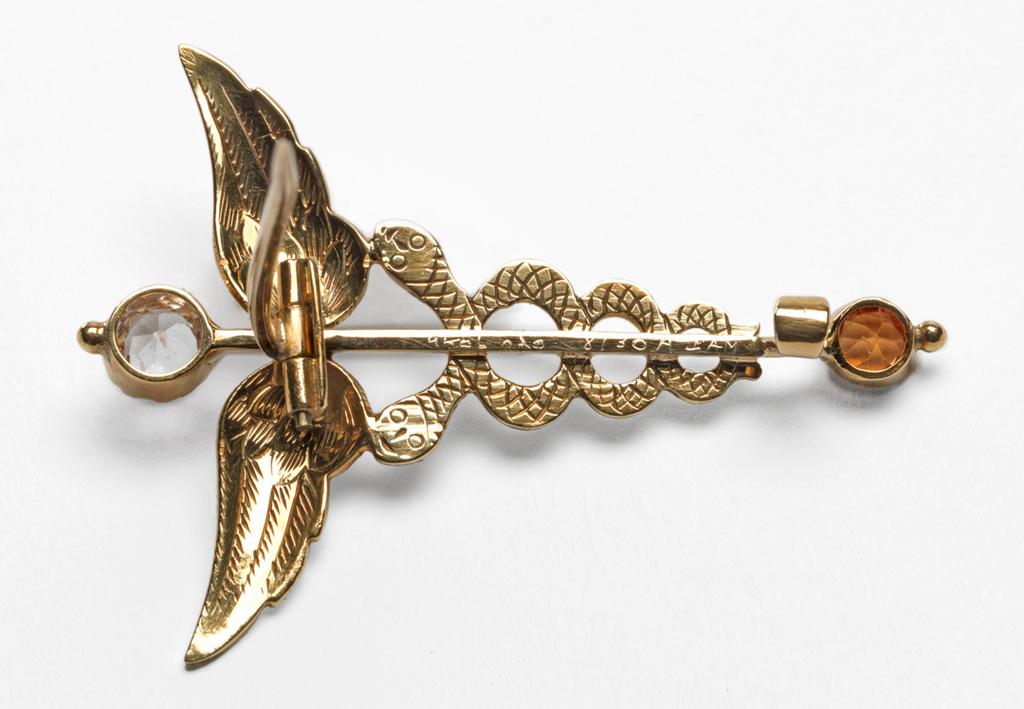 An image of Jewellery. Caduceus Brooch. Giuliano, Ferdinand, London. In original cream leather case (A), the silk lining of the lid printed in black with ‘BEATRICE CAMERON/102, MOUNT STREET, W/Art Jewellery by FERDINAND GIULIANO’. Gold, engraved, enamelled in red, green, and white, and set with a white sapphire and a citrine, height, whole, 4.7 cm, width, whole, 3.4 cm, circa 1910, pobably after 1903. Acquisition Credit: Given by Mrs J. Hull Grundy.