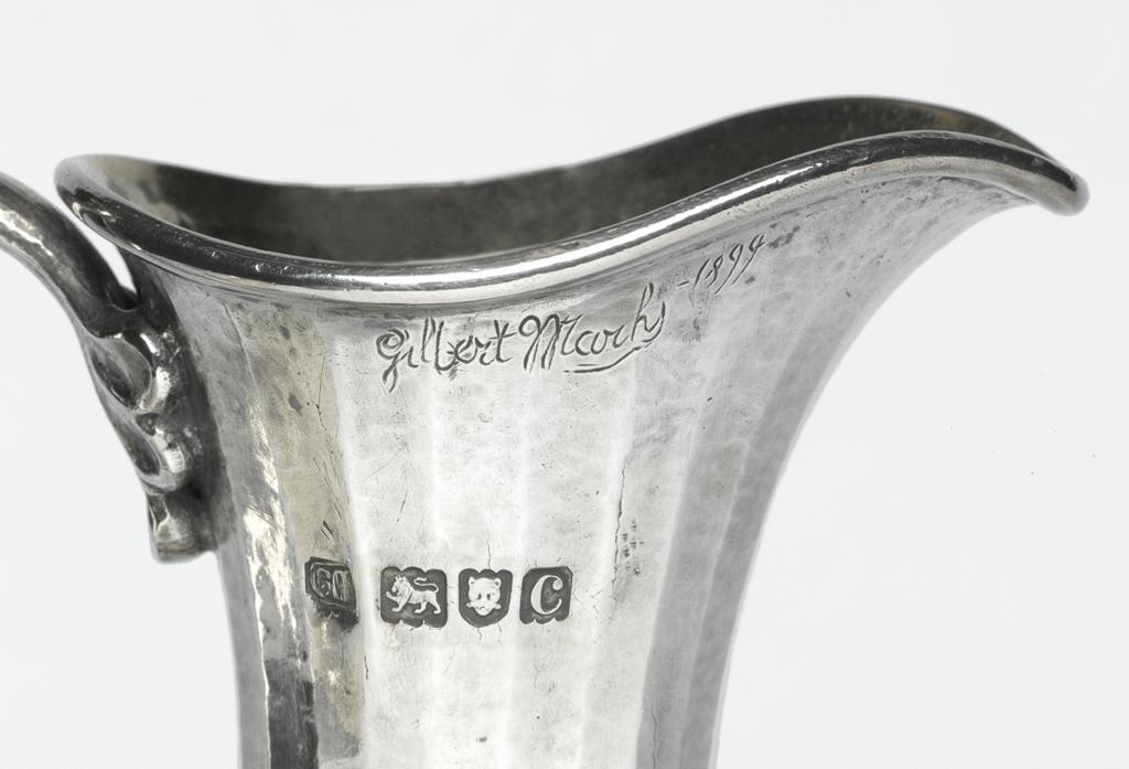 An image of Claret jug. Marks, Gilbert Leigh, silversmith (1861-1905). Facetted, lightly hammered sides decorated with a swag of vine leaves, grapes and ribbons. The tall slender jug has an ovoid body, which is facetted and spot hammered, and is decorated round its shoulders with a swag of vine leaves, grapes on ribbons. It stands on a slightly domed circular food, which is embossed with a border of stylised leaves. The cast harp-shaped handle has stylised shell terminals. Silver, facetted and spot-hammered, with embossed and chased decoration, and cast handle; the interior gilt, height, to top of handle, 30.6 cm, width, overall, 15.4 cm, weight, whole, 860 g, 1898-1899. Arts and Crafts.