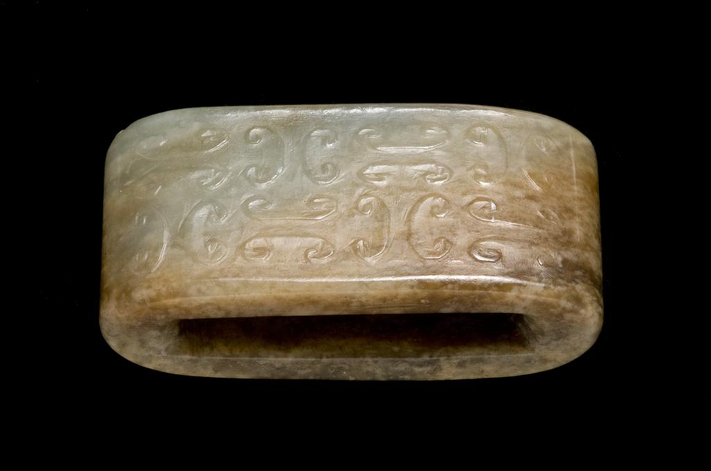 An image of Scabbard Slide. Grey-green jade with large areas of brown staining. One side decorated with C-shaped volutes and the other with rows of small spirals in relief. Nephrite, height 1.45 cm, length 4.6 cm, width 2.2 cm, 1368-1644 AD, Chinese.