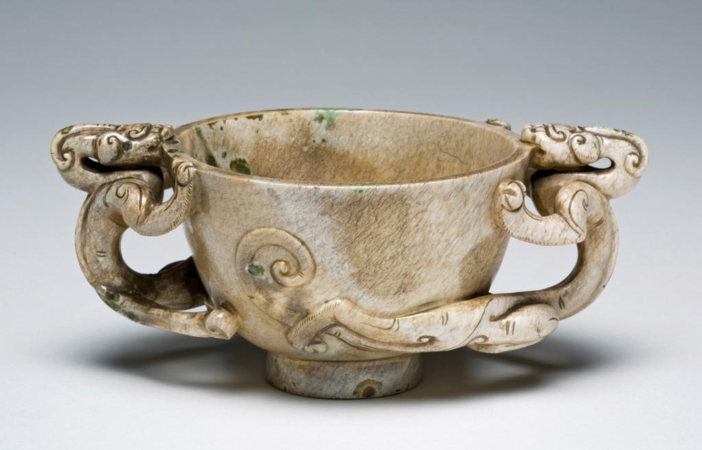 An image of Cup/ Greyish jade cup resting on a tall footring. The handles are decorated with feline dragons in high relief. Incised on the footring the character yong, eternal. Jade cups in this form are very commonly seen in the late Ming dynasty. It is possible that this object has been treated to give the appearance of having been buried and of great age. Nephrite, height 6 cm, width 14.7 cm, 1500-1700. Chinese.