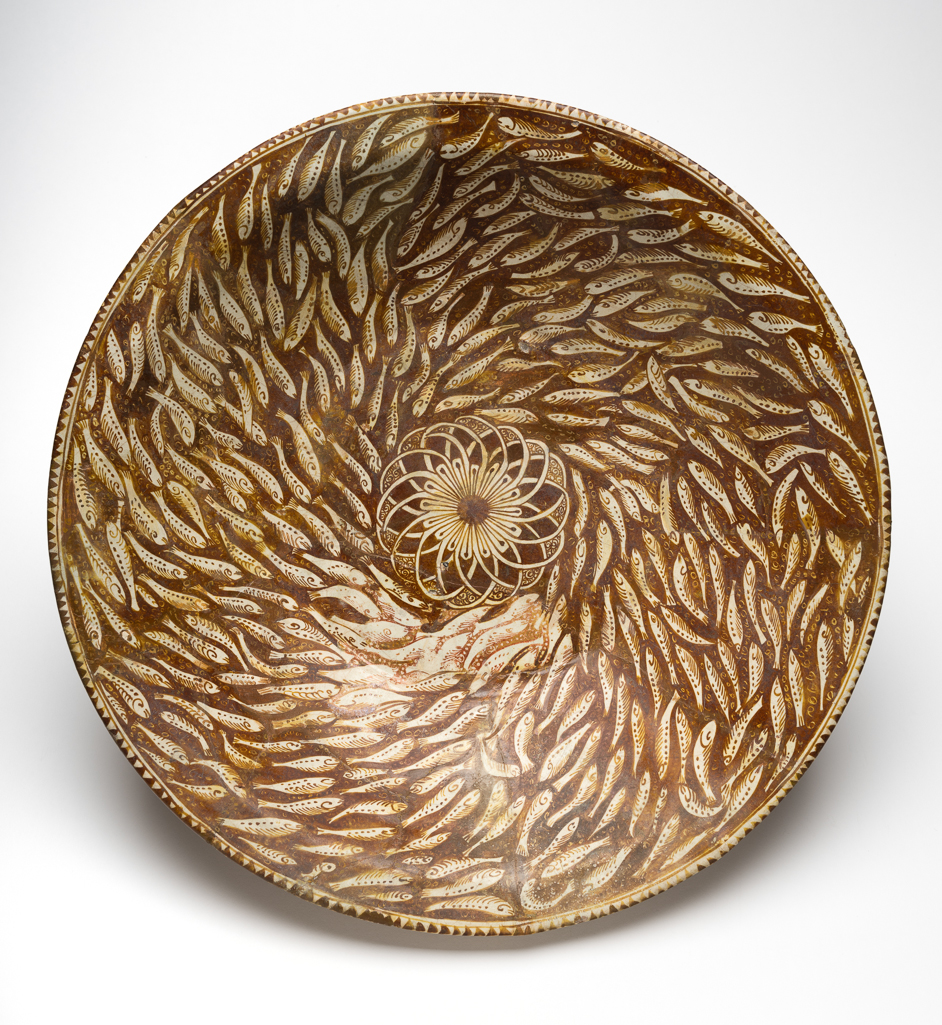 An image of Islamic Pottery. Dish. Unknown potter, Iran, probably Kashan. Shape: conical bowl with a low foot ring and a plain rim. Interior: on the rim a dentillated pattern is painted in lustre. A dense shoal of fish is painted on the body, swimming in a whorl around the roundel which contains a rosette with overlapping petals. Both fish and rosette are painted in reserve against a lustred background into which scrolls are incised on the body. Exterior: a dentillated pattern, of larger scale than that on the interior, is painted on the rim. On the body ‘ear muff’ style palmettes are painted in reserve against a lustred background, below which a pair of lustre concentric lines mark the edge of decoration. Glaze runs unevenly onto the foot ring. Pinkish fritware, wheel thrown and painted in brown pigment and lustre under or over a white glaze, height, whole, 12 cm, width, whole, 45.7 cm, diameter, base, 22.8 cm, circa 1260-circa 1350. Il-Khanid. Notes: Possible that a large section of the decoration was added in recent times.