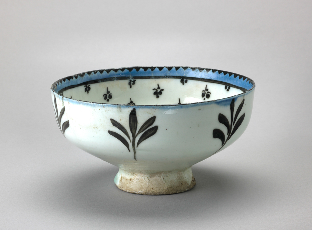 An image of Islamic pottery. Bowl. Production Place: Kashan. Shape: carinated bowl with a plain rim, sits on a slightly flared foot ring. Interior: on the rim black dentilation is reserved in a band of blue that is enclosed by a black line. On the body, a fleurs-de-lys pattern radiates from the base. In the roundel, framed by a black band and dashes, a stylised floral spray is reserved in blue. Glaze defects on the roundel. Exterior: on the rim blue pigment spills over from the interior. On the body black water weeds are painted. Glaze covers the surface, running unevenly on the foot ring; a light covering of glaze is present on the underside of the foot ring. Fritware, probably wheel thrown, painted in black and blue under a transparent white glaze. 1200-1220. Lent by the Ades family.