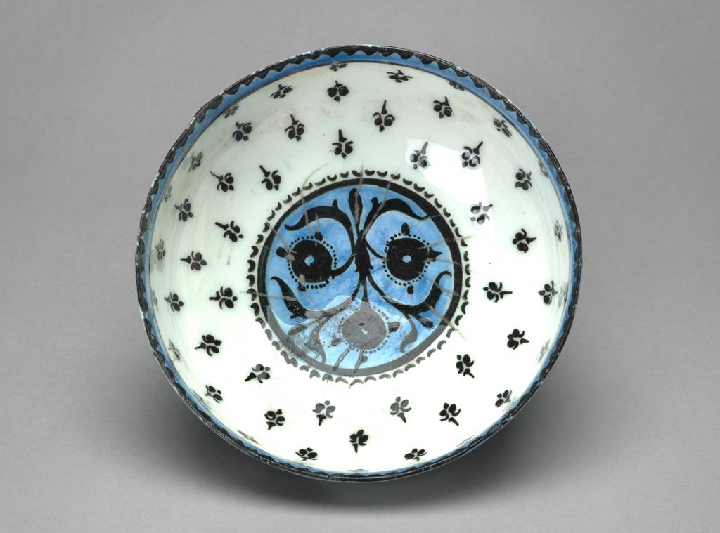 An image of Islamic pottery. Bowl. Production Place: Kashan. Shape: carinated bowl with a plain rim, sits on a slightly flared foot ring. Interior: on the rim black dentilation is reserved in a band of blue that is enclosed by a black line. On the body, a fleurs-de-lys pattern radiates from the base. In the roundel, framed by a black band and dashes, a stylised floral spray is reserved in blue. Glaze defects on the roundel. Exterior: on the rim blue pigment spills over from the interior. On the body black water weeds are painted. Glaze covers the surface, running unevenly on the foot ring; a light covering of glaze is present on the underside of the foot ring. Fritware, probably wheel thrown, painted in black and blue under a transparent white glaze. 1200-1220. Lent by the Ades family.