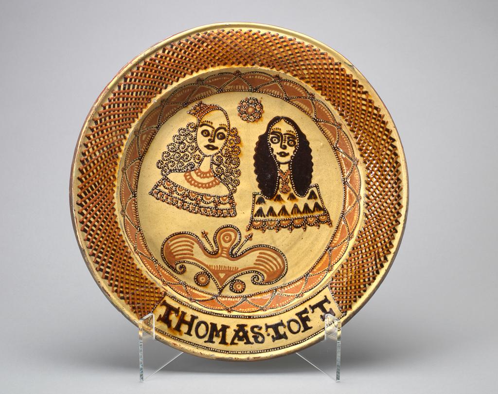 An image of Dish/Charger. Charles II and Catherine of Braganza. Toft, Thomas (British, act.1663-1689). Production Place: Staffordshire. Circular with a broad rim, deep sides, and almost flat centre. Decorated in the middle with busts of a woman with her hair dressed in ringlets and wearing a necklace and a crown, and a man with a long wig, probably Catherine of Braganza and Charles I. Above and between their heads is a rosette, and below, a winged motif. The sides of the well are decorated with a zig-zag slightly curved line forming triangles, and the rim has a trellis border, broken at the bottom by a rectangular panel enclosing the name THOMAS.TOFT. Earthenware, thrown, the front coated with white slip, and slip-trailed in two shades of brown under lead-glaze; reverse unglazed, diameter, whole, 42.5 cm, circa 1662-circa 1685.