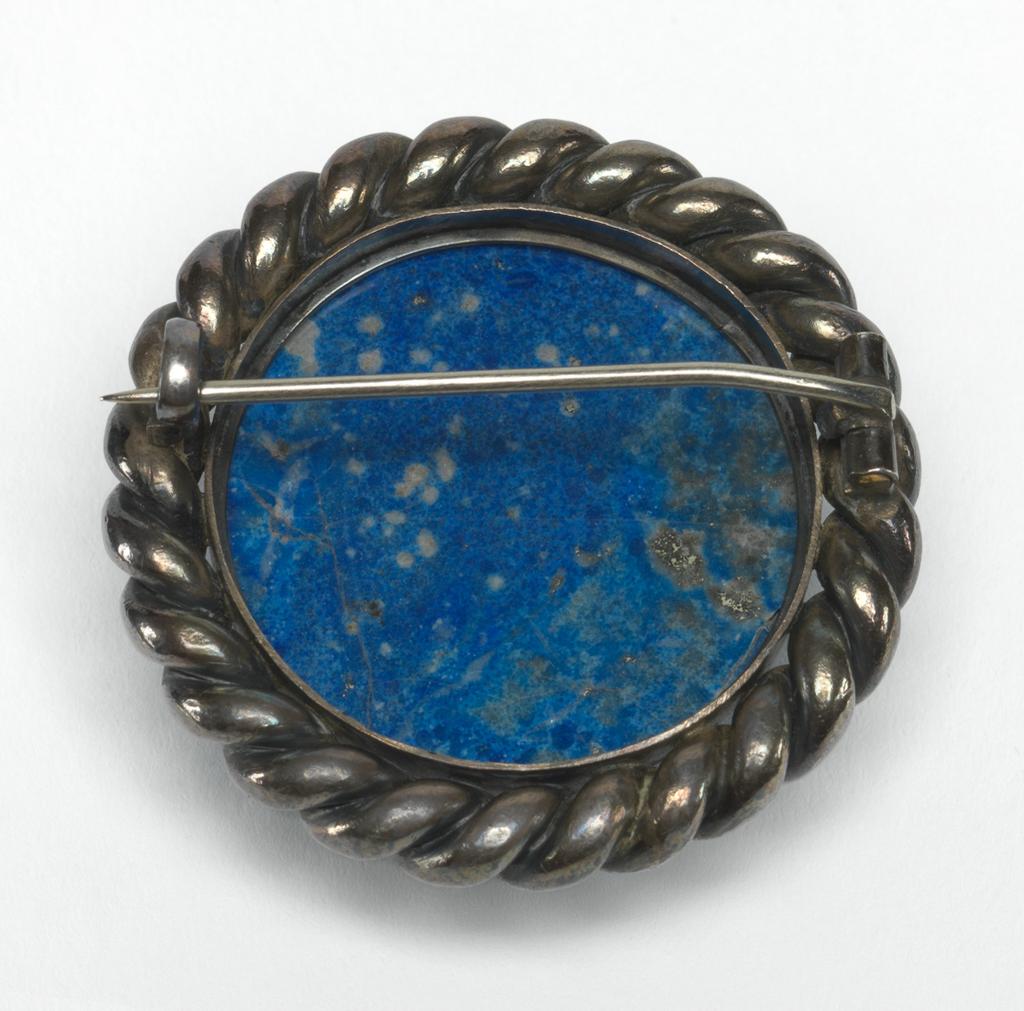 An image of Lapis lazuli and silver brooch. Stein, Michael (possibly); Stein, Leo probably (1872-1947). Cabochon lapis lazuli with pyrite and calcite inclusions, in a circular silver setting with a twisted 'rope' border; pin fastening across the back, diameter 4.2 cm, circa 1900. Made in Colmar, Paris.