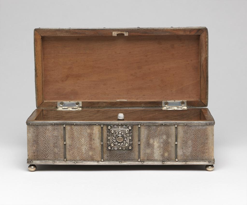 An image of Box/objet de vertu. Cooper, John Paul (British, 1869-1933). Colarossi, Lorenzo, maker of box (1879-1965). Rectangular with four silver bun feet, a button-operated fastener at centre front and a slightly convex lid attached to the lower part by two silver hinges. The sides and lid are divided into five panels by strips of silver with silver studs at regular intervals, and the edges are mounted in silver with rope edging. The button of the clasp is surrounded by a square silver plate embossed with a wreath of curling leaves and four studs, and has a scalloped outer edge with four large and four small studs. Walnut covered in rough shagreen, mounted in silver with a silver clasp, height, overall, 8.6 cm, length, overall, 22 cm, width, overall, 10.2 cm, circa 1932. Arts and Crafts.