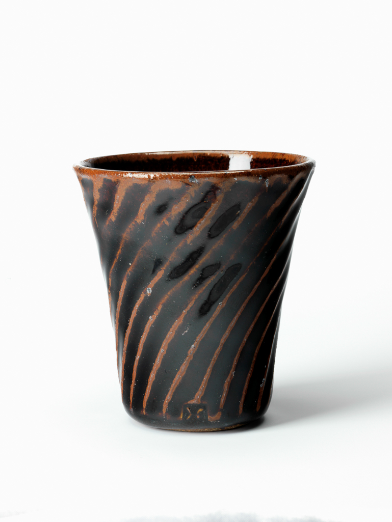 An image of Studio Ceramics. Beaker. Hughes, Edward (British, 1953-2006). Of truncated conical form with wrythen fluting on the exterior. Buff stoneware, thrown, carved with wrythen fluting, and covered with dark brown tenmoku glaze, probably over iron-bearing slip; base unglazed. Height, whole, 8.8 cm, diameter, rim, 8.1 cm, circa 1988-1989. In 1989 Hughes moved his workshop from Renwick to Isel Hall, Cumbria.
