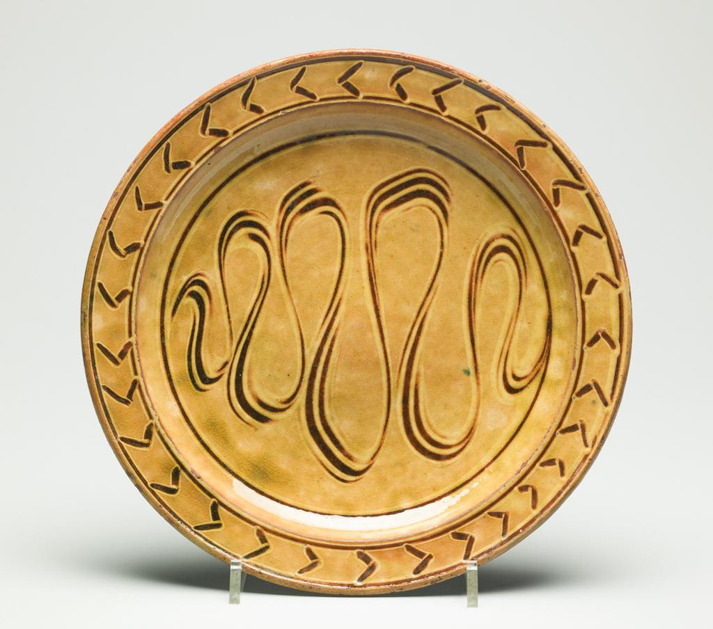 An image of Studio Ceramics. Plate. Cardew, Michael (British, 1901-1983). Galena glazed earthenware slipware plate with raised rim. Combed meander decoration in well, with chevrons around the rim. Height 3.2 mm, diameter, base, 14  mm, diameter, widest point, 26.9 mm, after 1929. Acquisition: Dr John Shakeshaft Bequest.