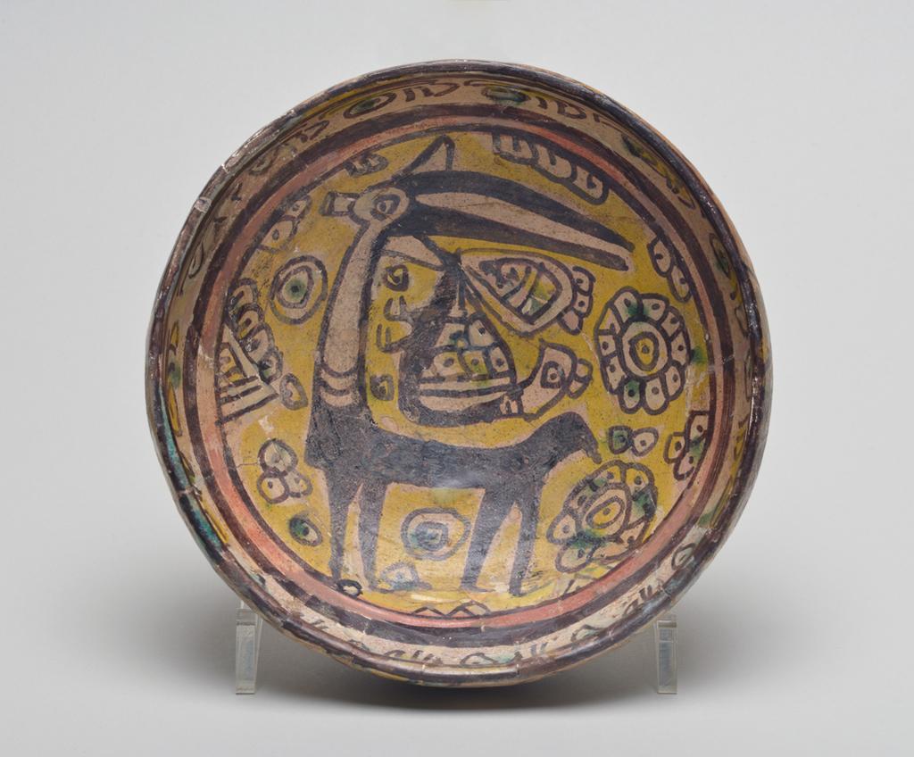 An image of Studio Ceramics.' Buffware' earthenware footed bowl. Underglaze painted in black and red slip, also yellow and green. Featuring gazelle, bird, flowers, and other motifs. Production Place: Nishapur, Iran. 900-999. Acquisition: Dr John Shakeshaft Bequest.