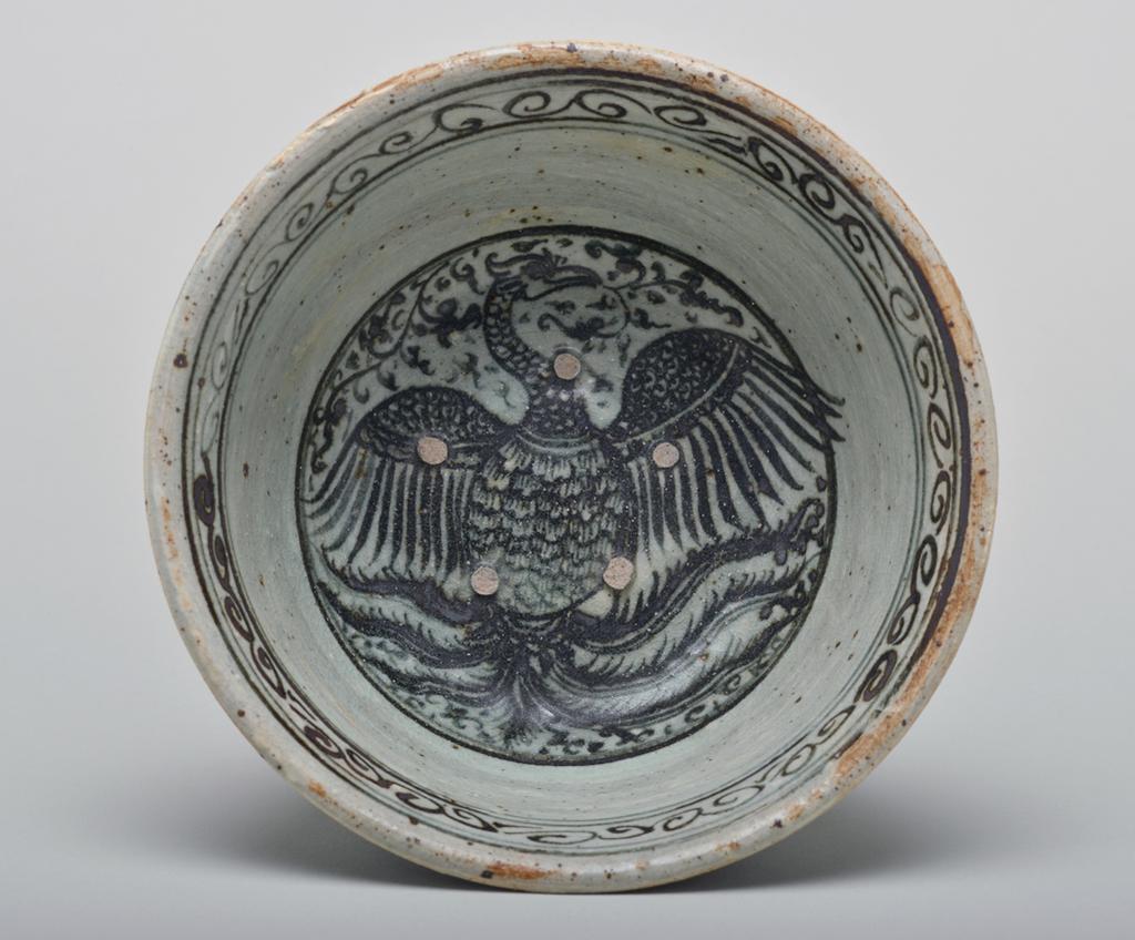An image of Studio Ceramics. Stoneware bowl with underglaze iron decoration featuring an unusual phoenix motif in a central medallion with foot. Production Place: Thailand. Light grey-green glaze with iron spots, pin-holing, and fine crazing. Five spur marks in the well. Signs of cylindrical kiln support on the base, indicating that this bowl was at the bottom of a stack. 1300-1399. Acquisition: Dr John Shakeshaft Bequest.