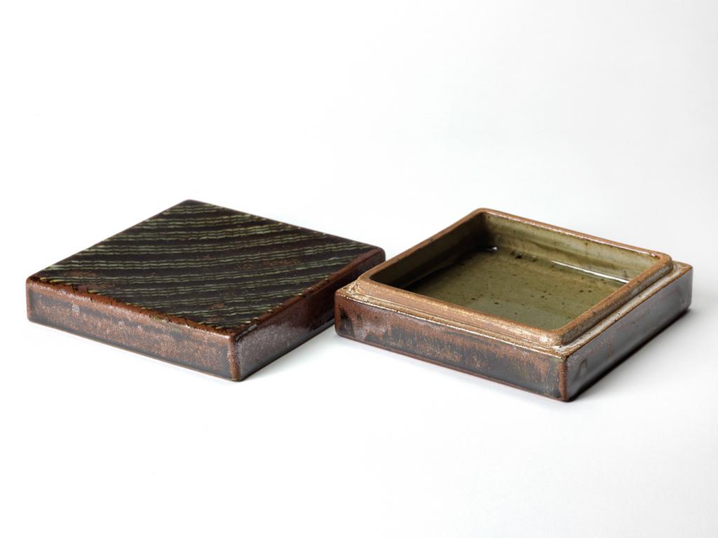 An image of Studio Ceramics. Box and cover. Hughes, Edward (British, 1953-2006). Square, with both halves of almost the same depth. Stoneware, slab-built in two parts, the interior coated with transparent, speckled grey-green glaze; the exterior with dark-brown and red-brown glaze; the cover trailed with diagonal stripes, and feathered. The base, the lower edge of the sides of the cover, and the upper edge of the sides of the box are unglazed. Height, whole, 6.2 cm ,width, base, 15.8 cm, length, base, 15.8 cm, 1989. Production Note: from the last firing at Renwick Pottery before the potter moved to Isel.