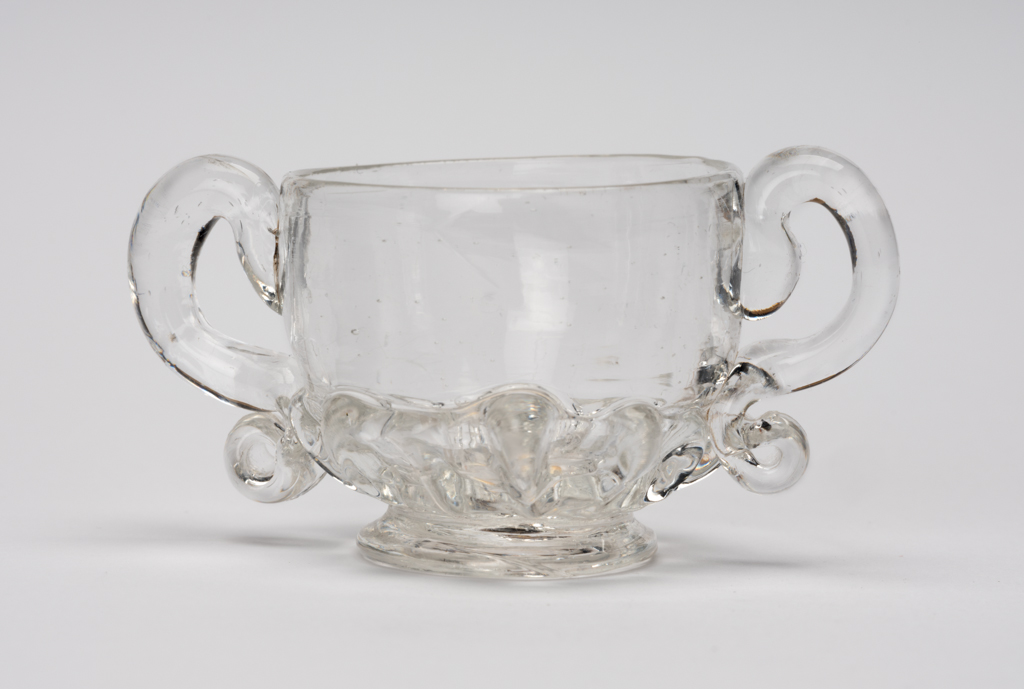An image of Miniature two-handled cup. Loving cup. Unidentified glasshouse, England. Clear lead-glass with gadrooned lower part and two S-shaped handles, height, whole, 3 cm, width, whole, 6.7 cm, 1690-1720. Baroque. Sir Ivor and Lady Batchelor Bequest through The Art Fund.