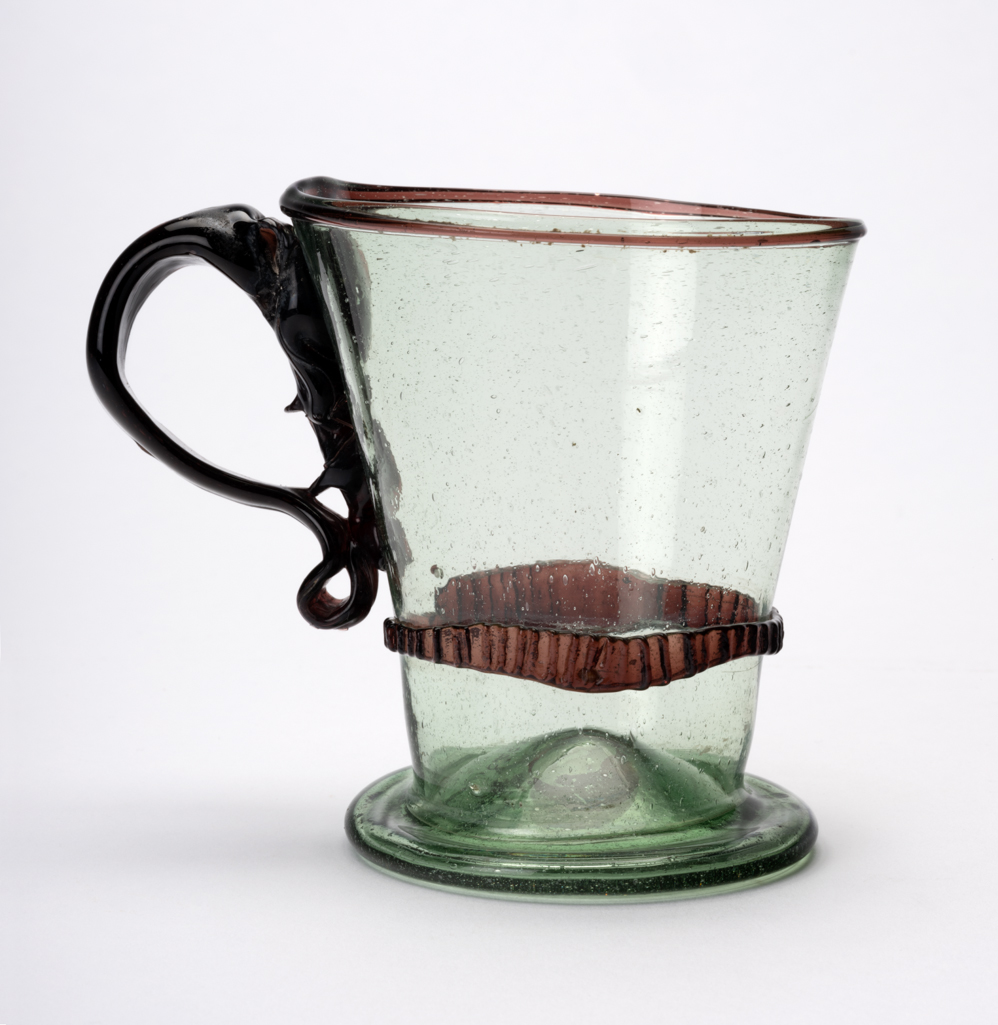 An image of Continental glass. Mug. Unknown glasshouse, Spain, Granada province. The mug has a projecting circular foot  with a pontil mark in the centre. The sides slope outwards to the rim which has an applied manganese-brown cord round its edge. A milled manganese-brown band of varying wide is applied a short distance above the foot. The handled is thicker and wider at the top than at its base which is curled under and extends upwards on the side of the mug to meet the top of the handle. Pale green glass, blown, with an applied manganese-brown cord, a milled brown band, and an applied brown handle. Height, whole, 10.4 cm, width, whole, 13.2 cm, diameter, rim, 9.3 cm, circa 1700-1800. Sir Ivor and Lady Batchelor Bequest.
