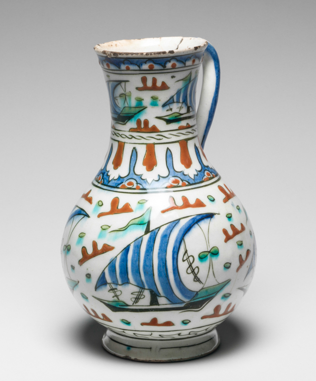 An image of Islamic pottery. Jug. Unknown potter, Turkey, Anatolia, Iznik. Shape: pear shaped body with slightly flared neck sitting on an out-turned foot ring. A strap handle with lower finger rest is attached just below the rim and on the shoulder. Exterior: just below the rim, a frieze of blue lobbed arches is outlined in black and contains a red central dot. On the neck and body, a frieze of lateen-rigged ships with blue and white stripped sails on black and green hulls and rigging float amongst red or green stylised islands, all outlined in black. On these ship friezes, the green pigment has run slightly into the white background. On the shoulder, separating the ship friezes, a band of dashed lines sits above lotus panel arcading in red and blue outlined in black. The handle is painted with blue horizontal and vertical stripes. On the lower body, a frieze of black scrolling lines is enclosed by sets of black concentric lines. Glaze covers the surface except the rim of the foot ring. Interior: surface is coated in glaze but otherwise undecorated. Fritware, probably thrown, coated in a white slip and painted in blue, red, green and black under a transparent white glaze. Height, whole, 22 cm, width, whole, 14 cm, diameter, rim, 8 cm, diameter, base, 8.5 cm, weight, whole, 562 g, circa 1570- circa 1590. Ottoman.