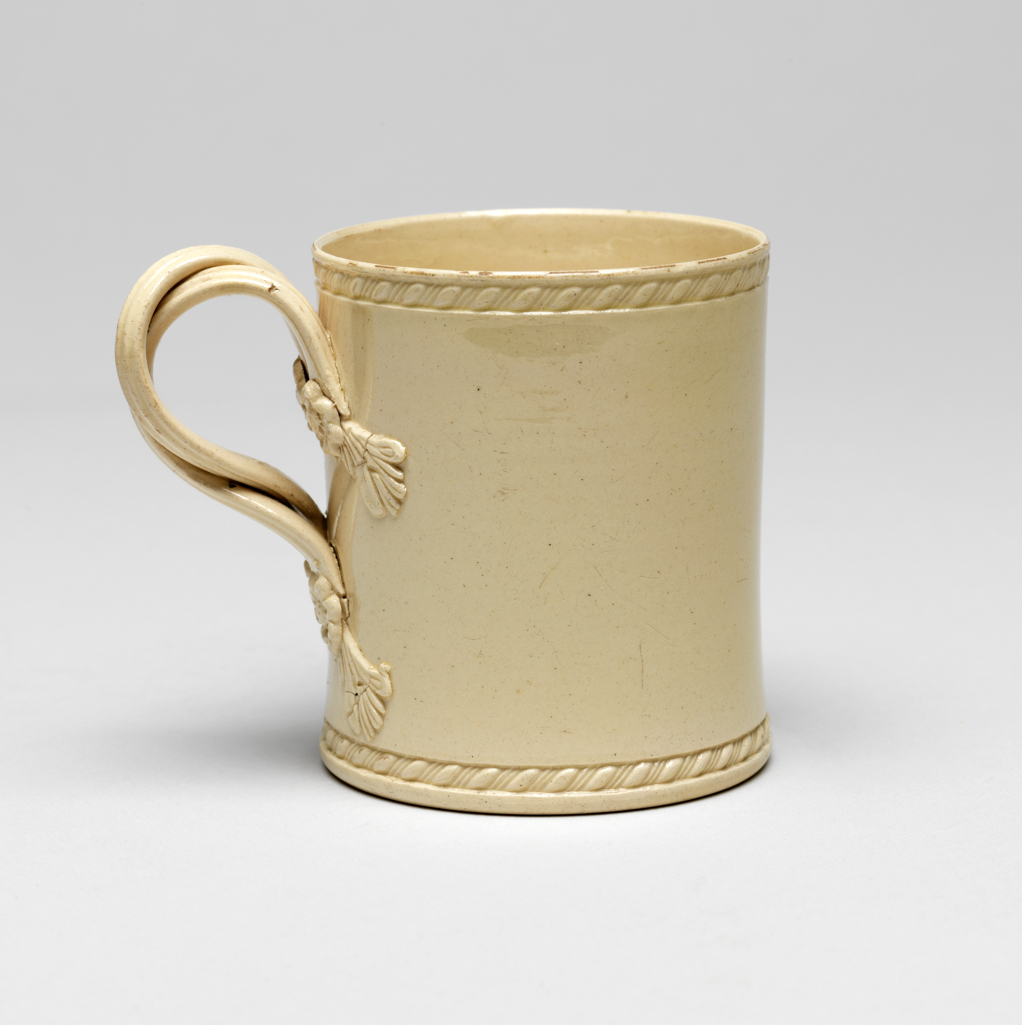 An image of Mug. Leeds Pottery, England. Cylindrical with a double interlaced reeded handle with moulded and applied flower and leaf terminals at the upper and lower ends. The upper and lower edges of the mug are rouletted with slanting oval beading. Deep cream-coloured earthenware with lead glaze, height, whole, 6.8 cm, diameter, base, 5.8 cm, width, whole, 8.4 cm, 1770-1780. Sir Ivor and Lady Batchelor Bequest.