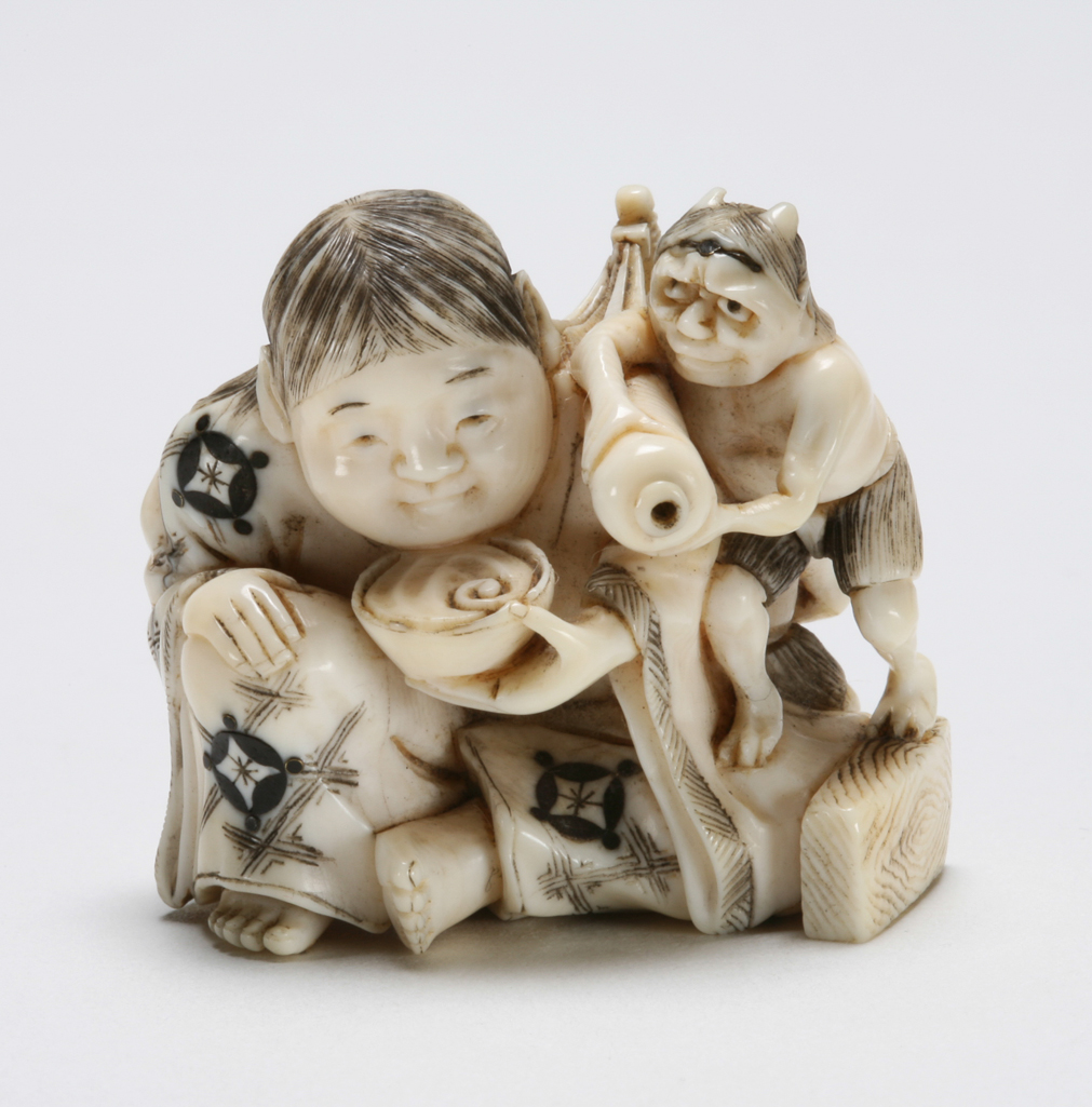 An image of Netsuke. Unknown maker, Japan. Shojo seated and drinking with two Oni attendants, one Oni serving Sake and sitting on the shoulder of the other Oni standing at the back, holding a large club. Ivory, carved, inlaid on costume and forehead of an Oni, in ebony, horn and copper alloy.1850-1900.