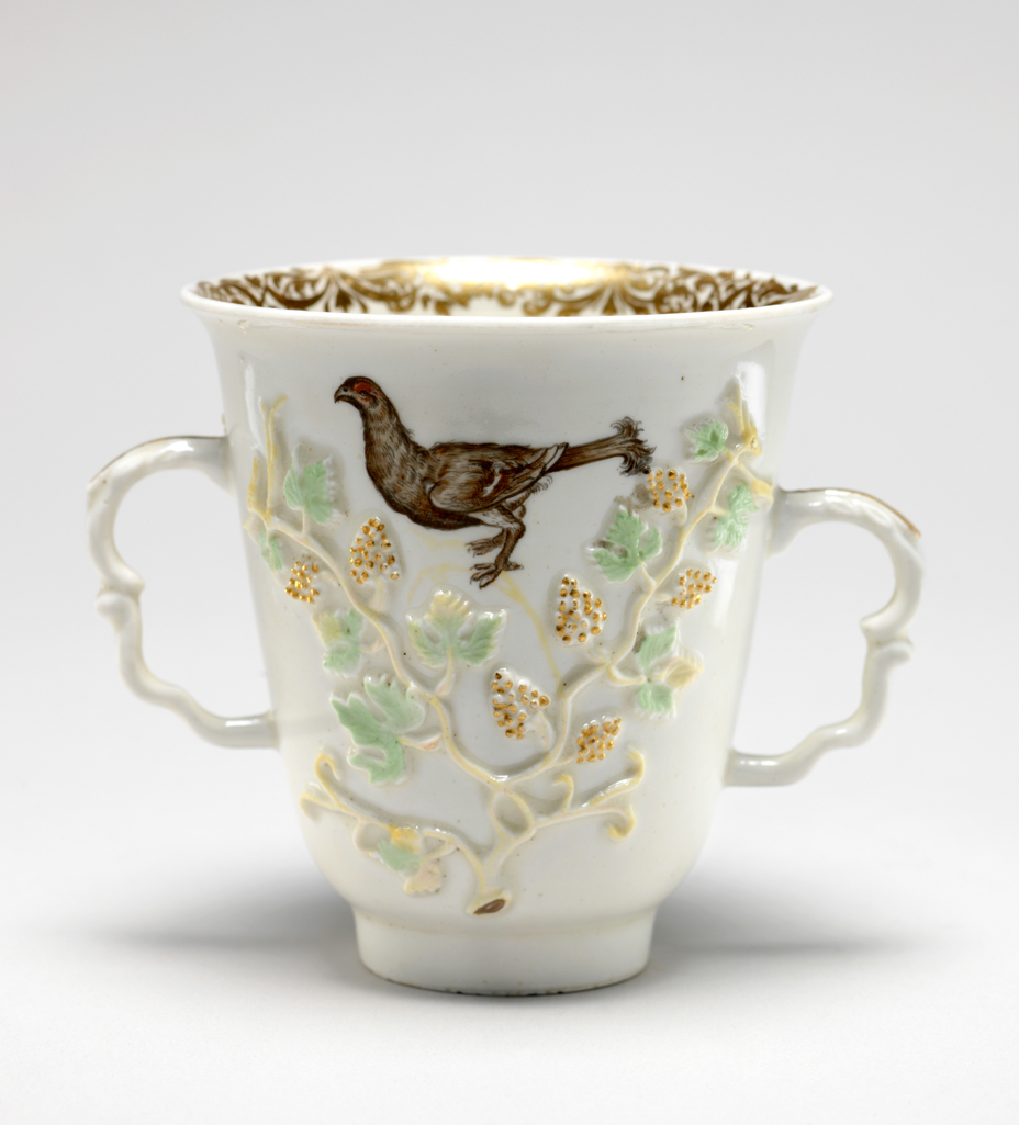 An image of Cup, with two scroll handles. Meissen Porcelain Factory, Germany. Applied decoration of pale green vine branches, and gilt grapes in relief. A black cock is painted on each side. On the inside of the lip is a band of floral scroll gilding. Hard-paste porcelain, gilding, height 8.3 cm, width, across handles, 10.2 cm, circa 1714-1720.