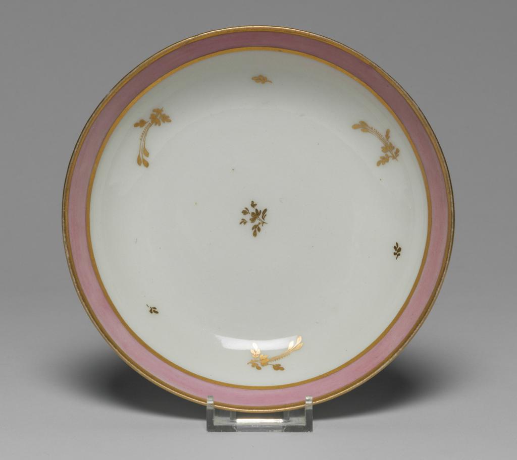 An image of Saucer. New Hall Porcelain Factory, Staffordshire. Hybrid hard-paste porcelain saucer, from a teaset. Decorated overglaze with a wide band of pink enamel between narrow gold bands and with scattered floral sprigs in gold. Pattern number 223. Height 3.2 cm, diameter 13.1 cm, 1785. See C.11A-L-1988: A service comprising a teapot and cover of 'silver' shape, a milk jug, sugar basin and cover, a slop basin, two plates of different sizes, two tea bowls, two coffee cups and two saucers.