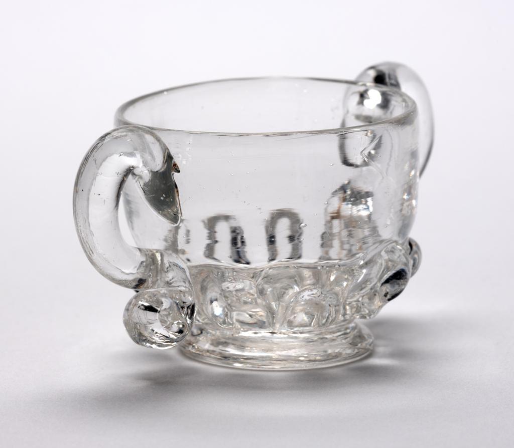 An image of Miniature two-handled cup. Loving cup. Unidentified glasshouse, England. Clear lead-glass with gadrooned lower part and two S-shaped handles, height, whole, 3 cm, width, whole, 6.7 cm, 1690-1720. Baroque. Sir Ivor and Lady Batchelor Bequest through The Art Fund.