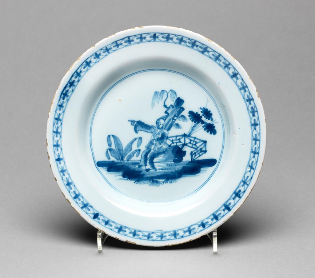 An image of English Delftware. Plate. Unidentified Liverpool pottery, England. Painted in blue with a Chinaman in a fenced garden, with flowering bamboo behind him. The circular plate has a sloping rim, shallow curved sides and flat centre. (Archer’s shape M but shallower) The well is decorated with a Chinaman seated on a rock and pointing to the viewer’s left with his raised right hand. Behind him there is a tall rock or tree trunk with plants growing from it, and a plant with three flowers projecting from a fence. To the left of the man there is another plant and in the foreground tufts of grass. The scene is surrounded by two narrow concentric circles. On the rim there is a narrow border of lozenge shapes with crosses over them. There are three peg marks on the reverse of the rim. Buff earthenware, tin-glazed bluish-white, and painted in blue. Height, whole, 23 cm, diameter, whole, 17.4 cm, circa 1760. Chinoiserie. Sir Ivor and Lady Batchelor Bequest through The Art Fund.