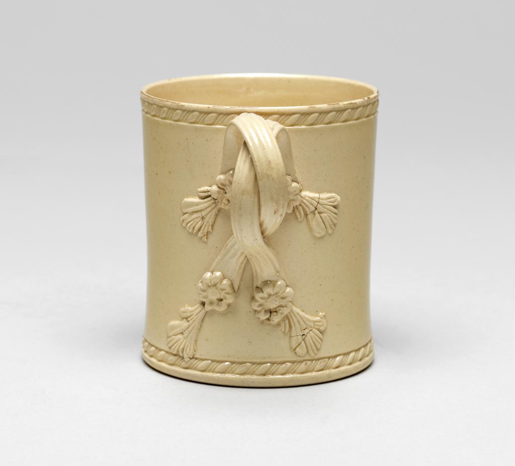 An image of Mug. Leeds Pottery, England. Cylindrical with a double interlaced reeded handle with moulded and applied flower and leaf terminals at the upper and lower ends. The upper and lower edges of the mug are rouletted with slanting oval beading. Deep cream-coloured earthenware with lead glaze, height, whole, 6.8 cm, diameter, base, 5.8 cm, width, whole, 8.4 cm, 1770-1780. Sir Ivor and Lady Batchelor Bequest.