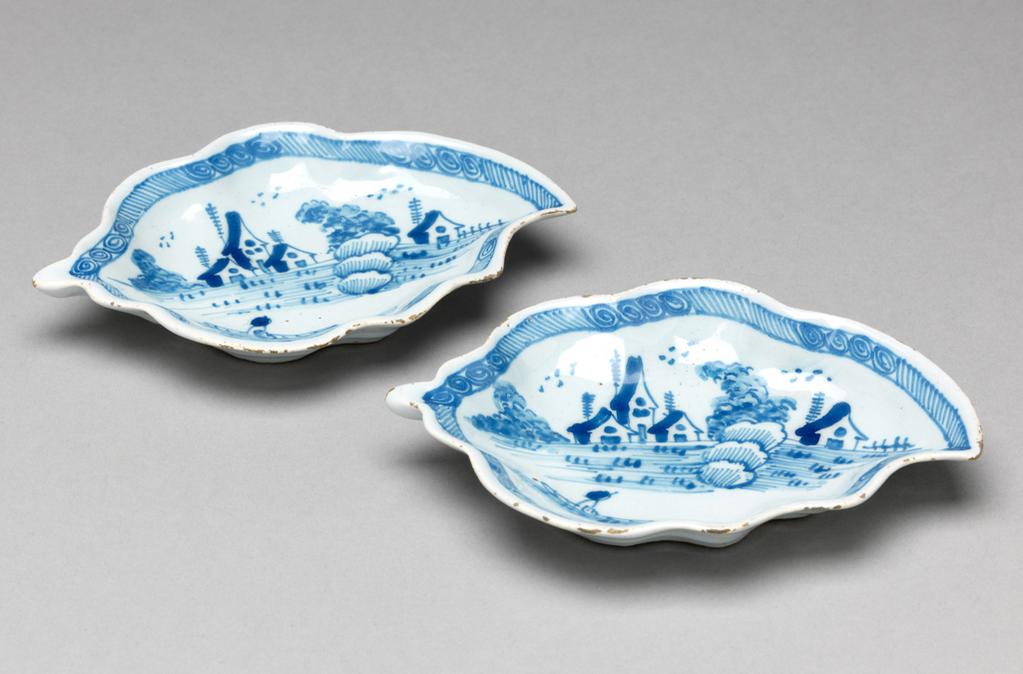 An image of English Delftware. Leaf dish. Pickle dish. Unidentified Liverpool pottery, England. Both dishes are moulded in the shape of a leaf with wavy edges and a short stalk, standing on a shaped footring. The interiors are painted with a lakeside scene in which there is a man in a boat in the foreground, an area with tufts of grass or reeds, three houses flanked by bushes and a single house with a fence to the right of it. Behind each house there is a slender tree resembling a telegraph pole, and overhead are three groups of schematic birds. The edge has a border of slanting lines broken by six groups of four spirals. Buff earthenware, press-moulded, tin-glazed pale bluish-white and painted in blue. Height, A, 2.2 cm, length, A, 13.2 cm, width, A, 9.2 cm; height, B, 2.2 cm, length, B, 13.1 cm, width, B, 9.3 cm, circa 1760. Rococo. Production Note: Small dishes of this shape were probably used for pickles presented on the table between the larger dishes in the course, or possible for sweetmeats for the dessert. Sir Ivor and Lady Batchelor Bequest through The Art Fund.