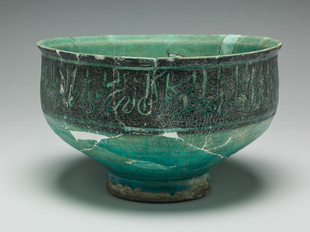 An image of Bowl. Islamic pottery. Silhouette ware. Unknown potter, Iran. Shape: irregular carinated bowl with conical lower body and cylindrical neck with an everted rim. Sits on a foot ring. Interior: on the body four black palmettes toped with finials are spaced at regular intervals, with an additional, smaller version at the centre of the base. Glaze pools in the base. Exterior: On the neck a wide band of black is incised with script with fleurs de lys at regular intervals. Glaze runs terminates on the foot ring, where coverage is uneven. Fritware, wheel thrown, partially coated in a black slip, carved under a transparent, crazed turquoise glaze. Height, whole, 12 cm, width, whole, 19.7 cm, diameter, base, 8.2 cm, weight 587 g, circa 1100-circa 1199. Seljuk.