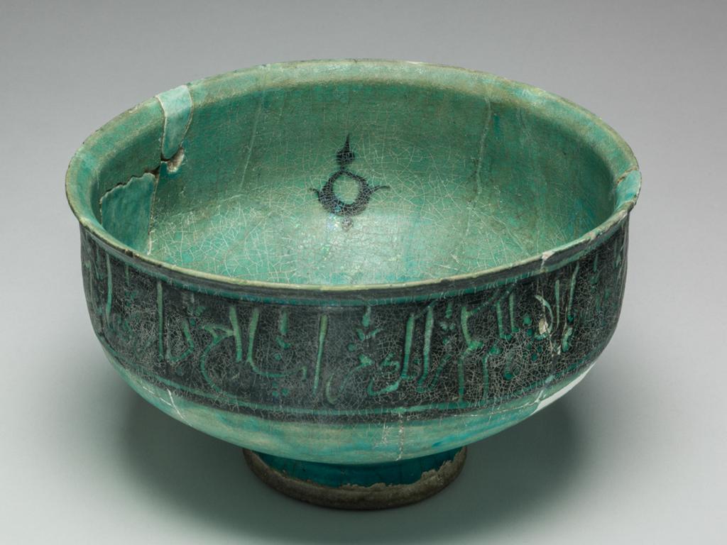 An image of Bowl. Islamic pottery. Silhouette ware. Unknown potter, Iran. Shape: irregular carinated bowl with conical lower body and cylindrical neck with an everted rim. Sits on a foot ring. Interior: on the body four black palmettes toped with finials are spaced at regular intervals, with an additional, smaller version at the centre of the base. Glaze pools in the base. Exterior: On the neck a wide band of black is incised with script with fleurs de lys at regular intervals. Glaze runs terminates on the foot ring, where coverage is uneven. Fritware, wheel thrown, partially coated in a black slip, carved under a transparent, crazed turquoise glaze. Height, whole, 12 cm, width, whole, 19.7 cm, diameter, base, 8.2 cm, weight 587 g, circa 1100-circa 1199. Seljuk.