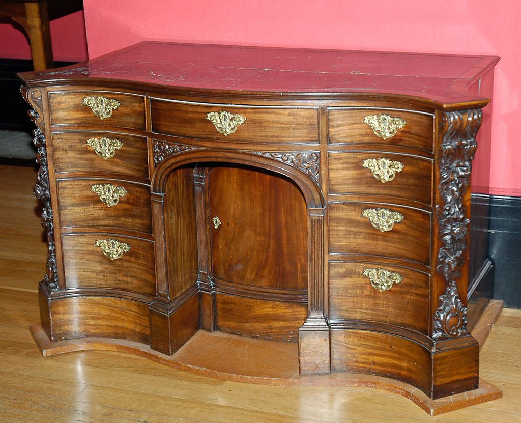 An image of Furniture. Kneehole desk/bureau dressing table. Mahogany kneehole desk with serpentine front, applied carving on the canted corners, leather inset top and ormolu mounts on the drawers. Kneehole and recessed cabinet framed with arch and fluted pilasters. Three of the drawers are frieze, while the recessed cupboard is flanked by six concave pedestal drawers. Mahogany, leather top, height 82.5 cm, width 142.4 cm, depth 72.5 cm, circa 1750. English.