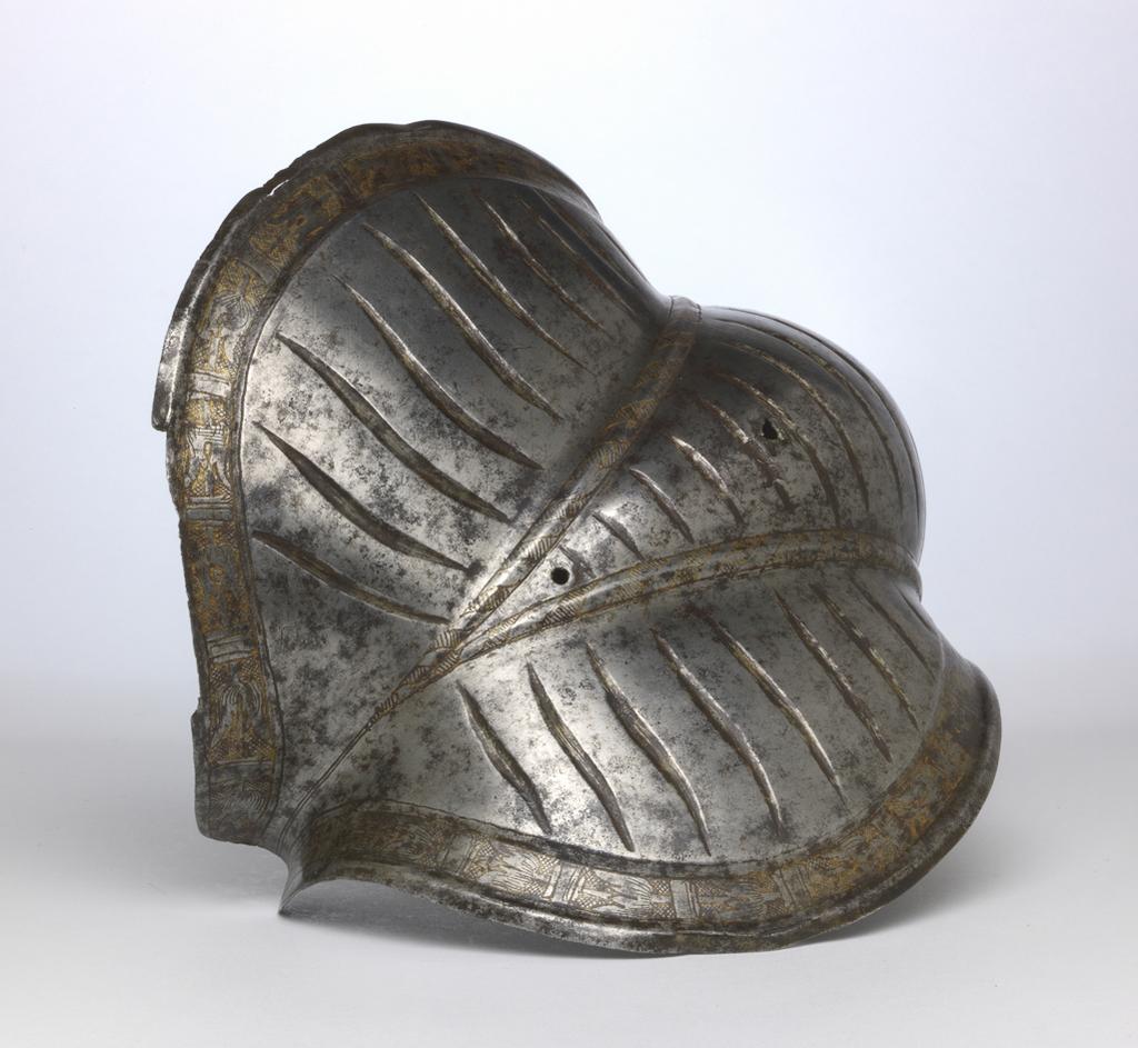 An image of Armour. Couter for the right elbow, for parade use, with 'puffed-and-slashed' decoration in imitation of civilian costume. Unknown production, South Germany. Formed of a single, shell-like plate, open at the rear and strongly shaped to the point of the elbow. Its outer edge is straight. Its upper and lower edges are convex and converge at the inside of the elbow in a strong, medial pucker. The upper and lower edges have partial inward turns accompanied by recessed borders decorated with etched and gilt tulip-like flower heads on a cross-hatched ground separated by broad bands enclosed by pairs of narrower bands. The boss over the elbow is enclosed by a pair of raised ribs that converge to the inside of the elbow. Each rib is decorated with etched diagonal bands that are alternatively hatched and gilt, and plain. The remainder of the couter, with the exception of its inner and outer ends is decorated with slender, vertical, recessed 'slashes' in imitation of those found on contemporary costume. The slashes on the upper and lower wings are slightly recurved, while those on the central boss are curved or straight. The front and rear of the central boss are each pierced with a rivet-hole for the attachment of the missing pair of vertical internal leathers that formerly connected the couter to the upper and lower cannons of the vambrace. The outer end of the couter is pierced with a pair of widely-spaced rivet-holes for the attachment of a missing bifurcated strap that terminated in a buckle to receive the missing strap that was formerly attached by a rivet occupying a now vacant hole located just to the inside of the medial pucker, and passed around the rear of the elbow. The couter is bright, with patches of dark patination. Its upper and lower edges both exhibit cracks. A small section at the inner end of the lower edge has broken away. The outer edge of the couter has a crack just below its centre, and its lower corner is bent inwards. Hhammered, shaped, riveted, wit