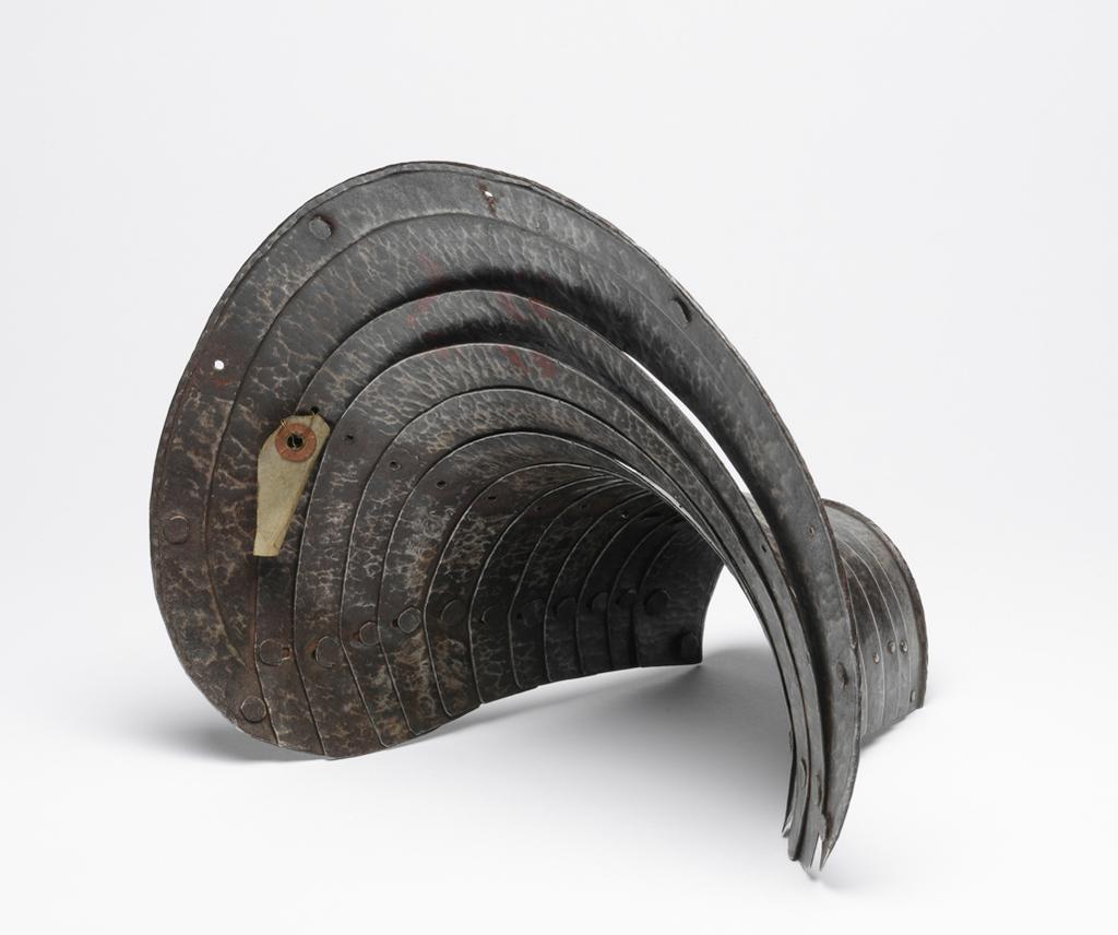 An image of M.9-1945: Armour. Defence for the left foreleg of a horse, for heavy cavalry use. Unknown production, Germany. Formed of twelve downward-overlapping lames curved to the front of the horse's upper foreleg and shoulder, and increasing in width to its upper end. The upper edge of the top lame is slightly convex with gently rounded corners. It has a file-roped inward turn accompanied by a recessed border containing nine holes for lining-rivets, of which six are occupied by externally-flush rivets. The lower edge of the bottom lame also has a file-roped inward turn. The outer ends of the lame on each are fitted with externally-flush rivets that formerly served to secure a strap around the rear of the horse's leg. The first to eleventh lames have bevelled outer edges. The lames are connected to one another at their outer ends by externally-flush sliding-rivets. The lames were further connected to one another by a pair of internal leathers located a short distance within the sliding-rivets. The leathers were attached to each lame by single externally-flush rivets. The rivets for the outer leather are preserved in the ninth to twelfth lames, and for the inner leather, in the eighth and ninth lames. The leg-defence has a ' black from the hammer' finish, now much worn, particularly at the centre of its lower half. Steel, hammered, shaped, riveted, with file-roped, recessed border and bevelled decoration. Height, whole, 25.5 cm, maximum, width, whole, 29.1 cm, maximum, depth, whole, 22.1 cm, maximum, weight, whole, 1.02 kg, circa 1550. Provenance: From the armoury of the Princes Radziwill, Castle of Niescwiez, Poland. Notes: forming a pair or near pair to M.14-1947. M.14-1947: Armour. Leg-defence for the right foreleg of a horse. Unknown production, Germany. Formed of twelve downward-overlapping lames curved to the front of the horse's upper foreleg and shoulder, and increasing in width to its upper end. The upper edge of the top lame is slightly convex with gently rounded c