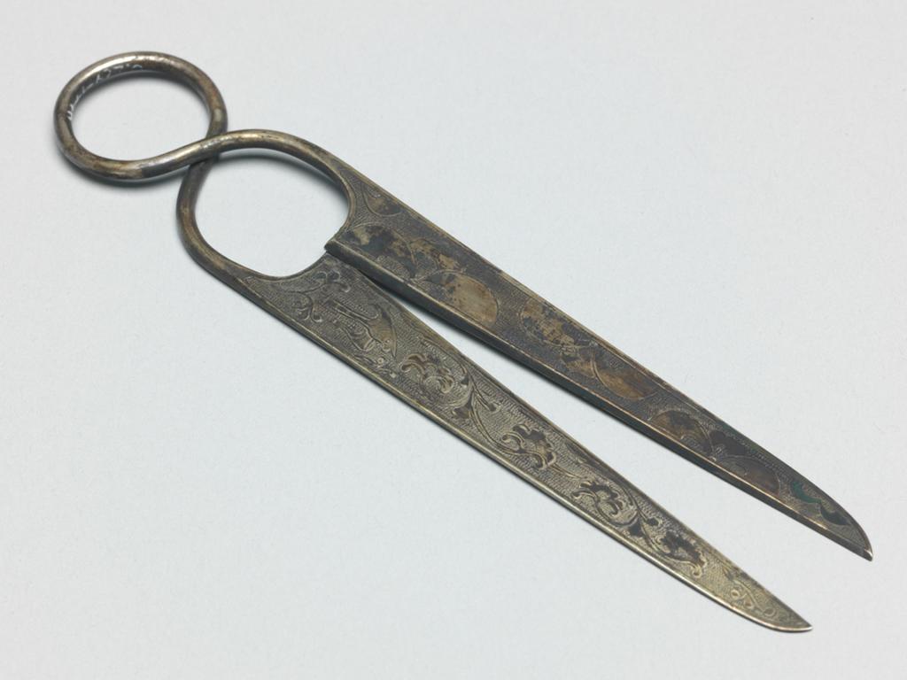 An image of Pair of scissors. Unknown maker, China. Pair of beaten silver alloy scissors. The handle is a wire spring shaped and the cutters are slightly convex on one side, and flat on the other. Each of the flat sides is decorated with a flying bird and a palmette scroll, and each of the convex sides with confronted palmettes. The pattern is bounded by a narrow plain border and chased on a ground of regular ring matting. Silver alloy, engraving, blades, chasing, length 14.9 cm, width 2.8 cm, T’ang Dynasty (618-906).
