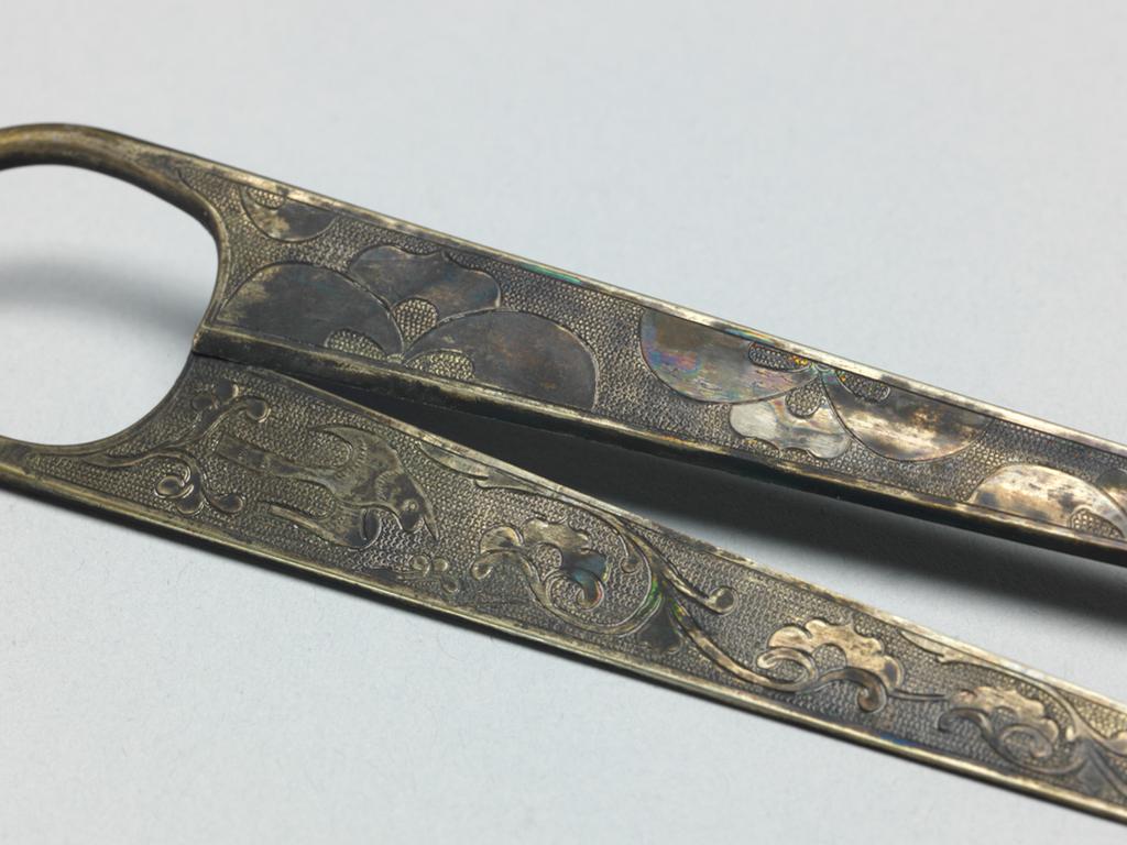 An image of Pair of scissors. Unknown maker, China. Pair of beaten silver alloy scissors. The handle is a wire spring shaped and the cutters are slightly convex on one side, and flat on the other. Each of the flat sides is decorated with a flying bird and a palmette scroll, and each of the convex sides with confronted palmettes. The pattern is bounded by a narrow plain border and chased on a ground of regular ring matting. Silver alloy, engraving, blades, chasing, length 14.9 cm, width 2.8 cm, T’ang Dynasty (618-906).