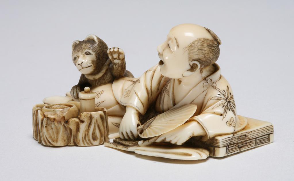 An image of Okimono. Unknown maker, Japan. A man reclining, resting his left arm on his books, while holding his pipe in his left hand and holding a fan in his right hand, he is pleased to see his pet cat on his legs, reaching out to the food container in the fore ground with its left paw. Ivory, 1870-1900.