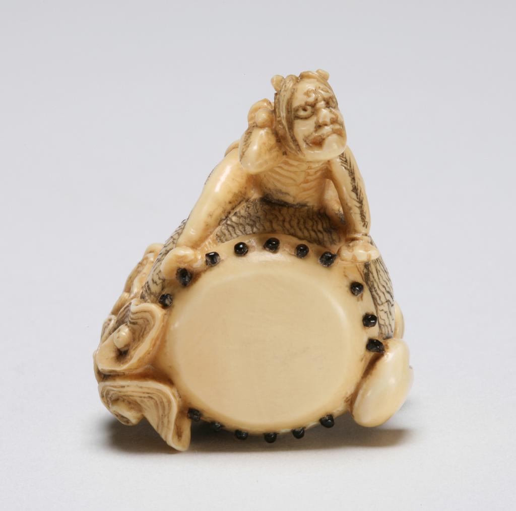 An image of Netsuke. Unknown maker, Japan. Raiden (God of thunder) drumming on a large Tai-go, that is elaborately decorated with clouds and some treasure objects like tobacco pouch with its Ojime and Netsuke. Plastic resin, the inlays are glass. 1970-2000.