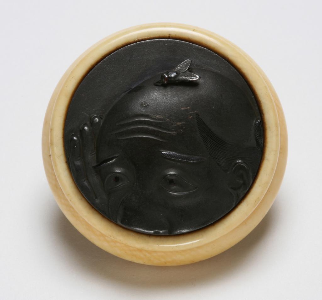 An image of Netsuke: kagamibuta. Unknown maker, Japan. Circular bowl, plain with indented ridge around the top edge and traces of green staining to inner rim, central himotoshi. Shibuichi disc, cast and decorated in relief with a fly resting on top of a man's shaved head above the wrinkled brow, the face cut off at the nose. The man's hand is raised as if to swat the fly. Finely stranded hair and eyebrows in shakudo and copper, the man's eyeballs in shakudo and copper, and the fly's eye is applied with copper. Ivory bowl with cast, metal disc decorated in relief and eyelet. Height, whole, 1.8 cm, width, whole, 4.2 cm, circa 1800-1868.