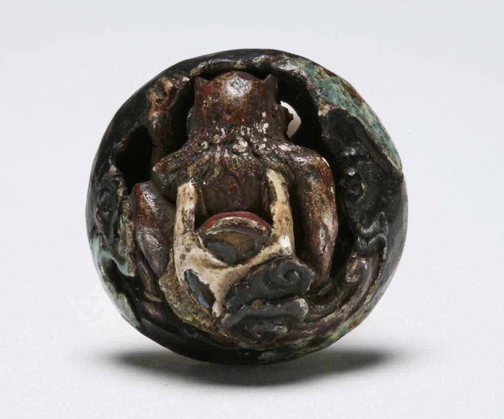 An image of Netsuke: katabori: Raiden the Thunder God with his drum. Unknown maker, Japan, Teiji or Sadaji. Raiden is sitting on his haunches with his knees up and his raised left hand holding on to the clouds; his drum is strapped to his back and he peers down through a hole in the clouds. Most of the glaze seems to have worn off, yet green clouds, a red band on the drum and white glazed eyes, back pack and shorts are still visible. There are possible traces of gold lacquer on his arms and the back of his head. Circular himotoshi just off centre. Earthenware and wood; circular with moulded relief and glazed, height, whole, 1.8 cm, width, whole, 3.4 cm, circa 1830-1870. Edo (Japanese period, 1615-1868). Notes(s): Raiden is the deity of thunder and storm. His destructive activities cause some fear but the farmers are grateful for the blessings the accompanying rain brings to the crops. Japanese artists give Raiden the appearance of a devil and often treat him with slightly humorous disrespect.