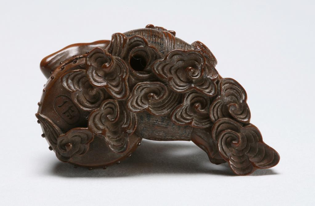 An image of Netsuke: katabori: Raiden seated with his drum. Masakazu, Edo Period, 19th century, Japan. Scowling, unshaven figure of Raiden seated on a cloud, his left arm resting on his thunder drum, his hand supporting his head; his right arm around his leg, his bag hanging at his waist. Fine incising and black ink details to tiger-skin loin cloth and leggings. His eyes are of ivory with dark pupils; his drum with two bands of raised, shiny black beading. Himotoshi at the rear of the drum and cloud. Wood, carved, incised with inalid decoration and ink detailing, height, whole, 3.1 cm, width, whole, 4.3 cm, circa 1800-1868. Edo Period (1615-1868).