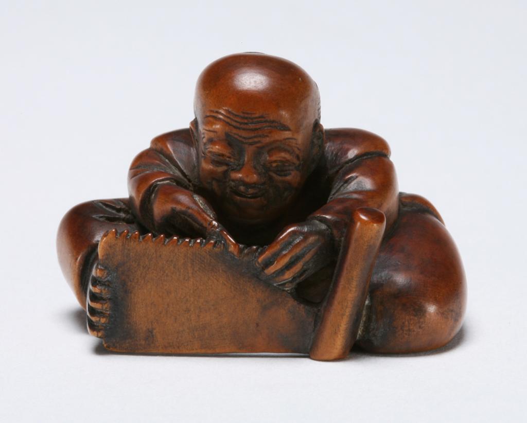 An image of Netsuke: katabori. Toolmaker. Unknown maker, Japan. Mitsutami, Edo Period (1603-1868), Japan. Seated figure of a toolmaker with a smiley, wrinkled face, sharpening the teeth of a saw which he steadies with one foot. Himotoshi on the underside of his leg alongside the mark. Boxwood, carved and stained, height, whole, 2.4 cm, width, whole, 3.7 cm, circa 1800-1868. Edo Period (1615-1868).